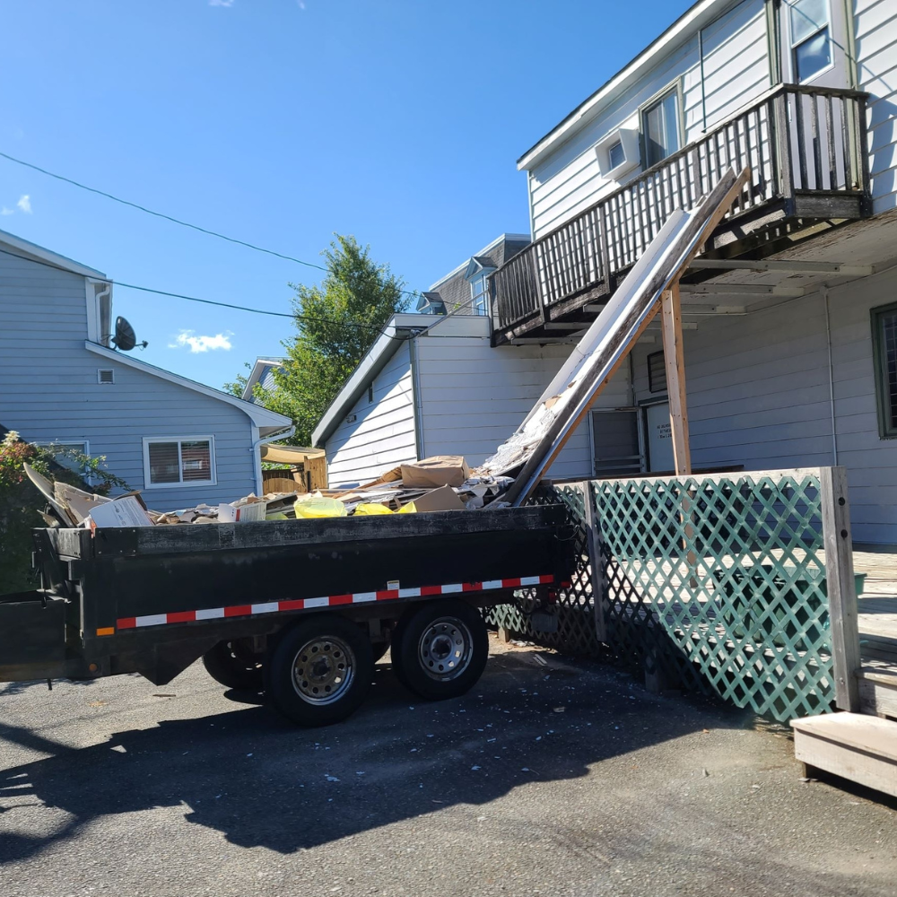 A black trash trailer filled with debris, with a temporary ramp leaning against the second floor of a white house with black railings on a sunny day.