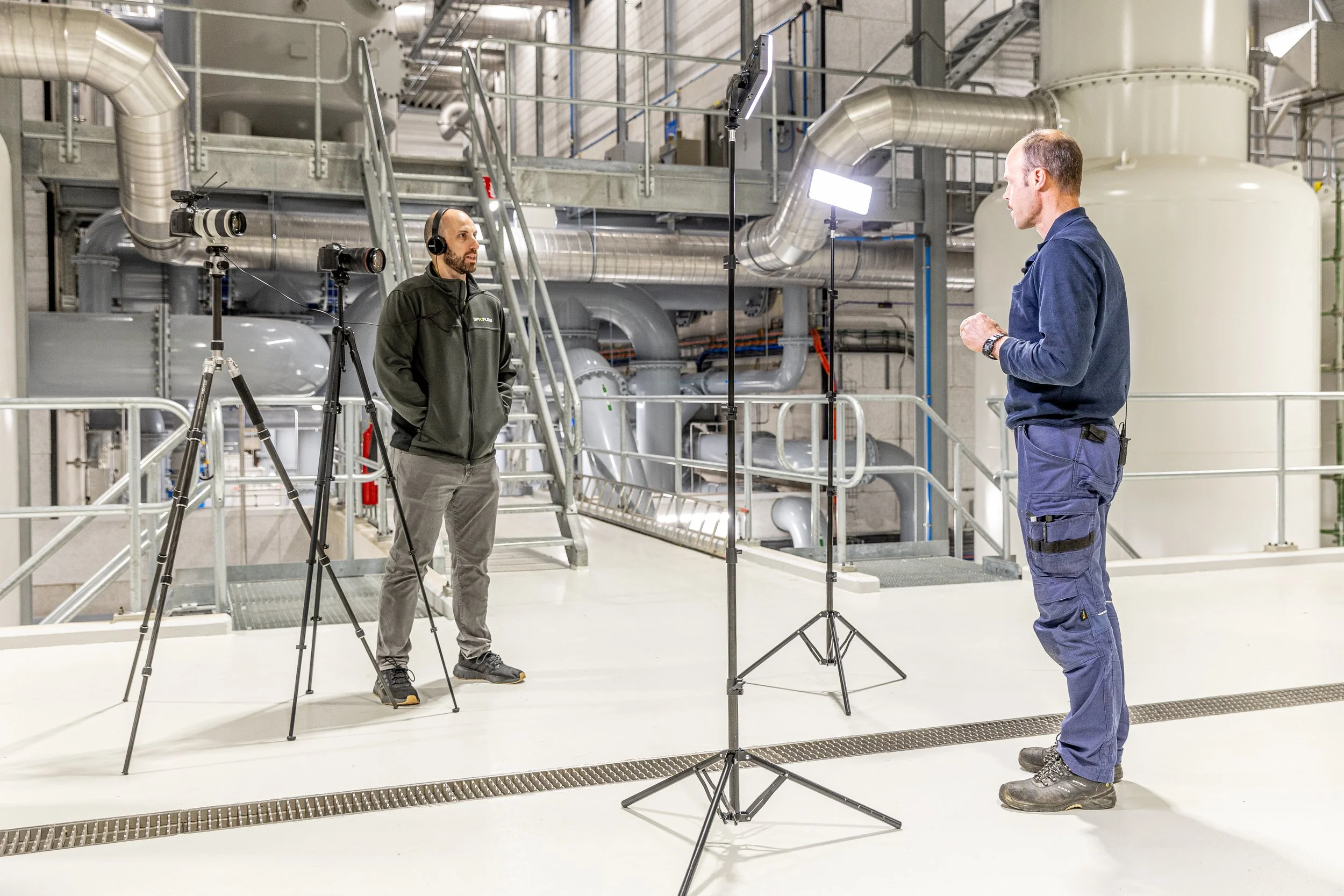 Two men in a factory setting are filming a video or taking photographs. One man, dressed in dark clothing and wearing headphones, stands with hands in pockets. The other man, in blue work clothes, is speaking or presenting. Lighting equipment and cameras are set up directed at the man in work clothes.