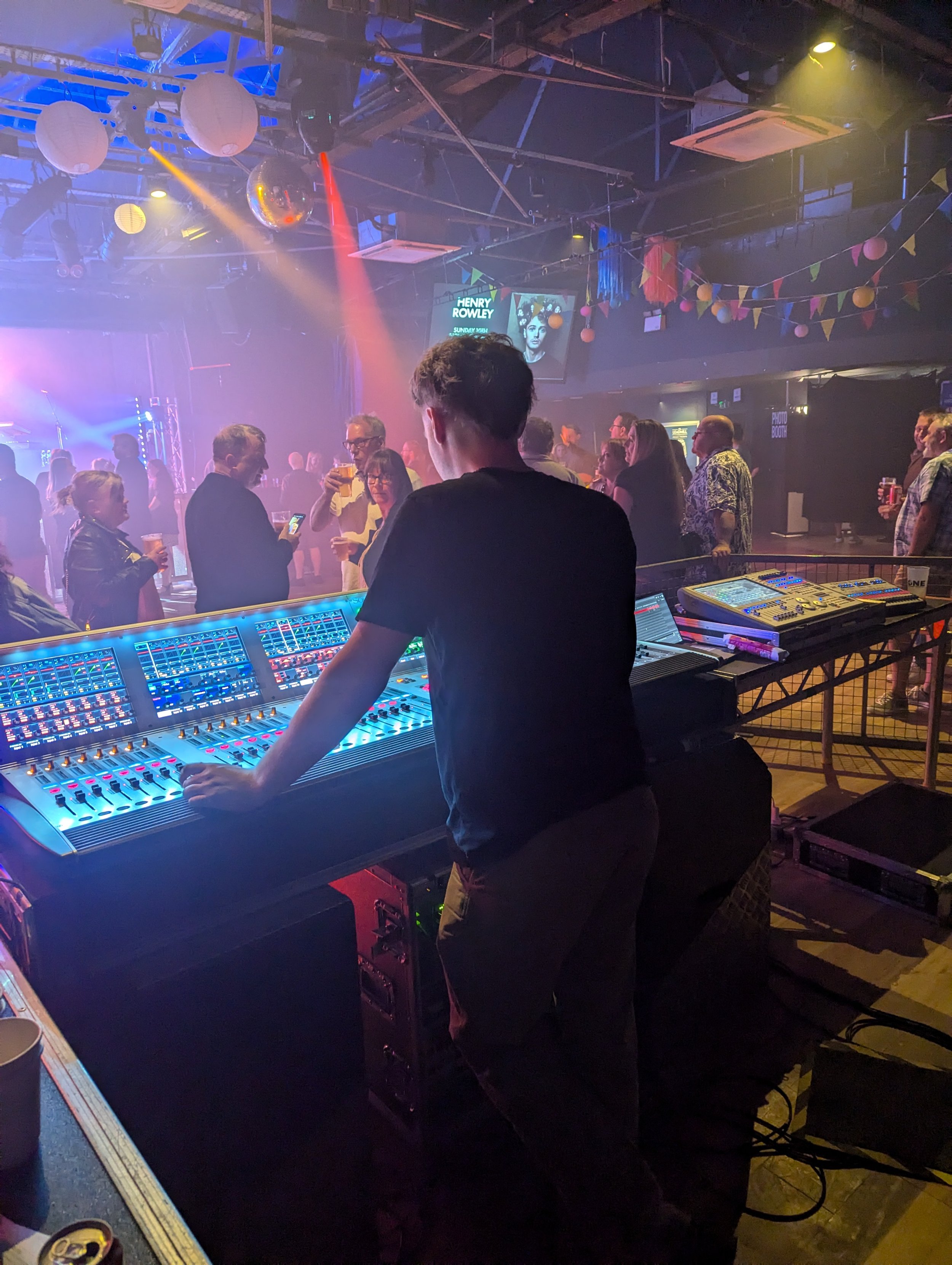 A DJ is playing music on a turntable at a night event with people dancing and drinking in the background. The scene is illuminated with colorful stage lights and decorated with paper lanterns and bunting.