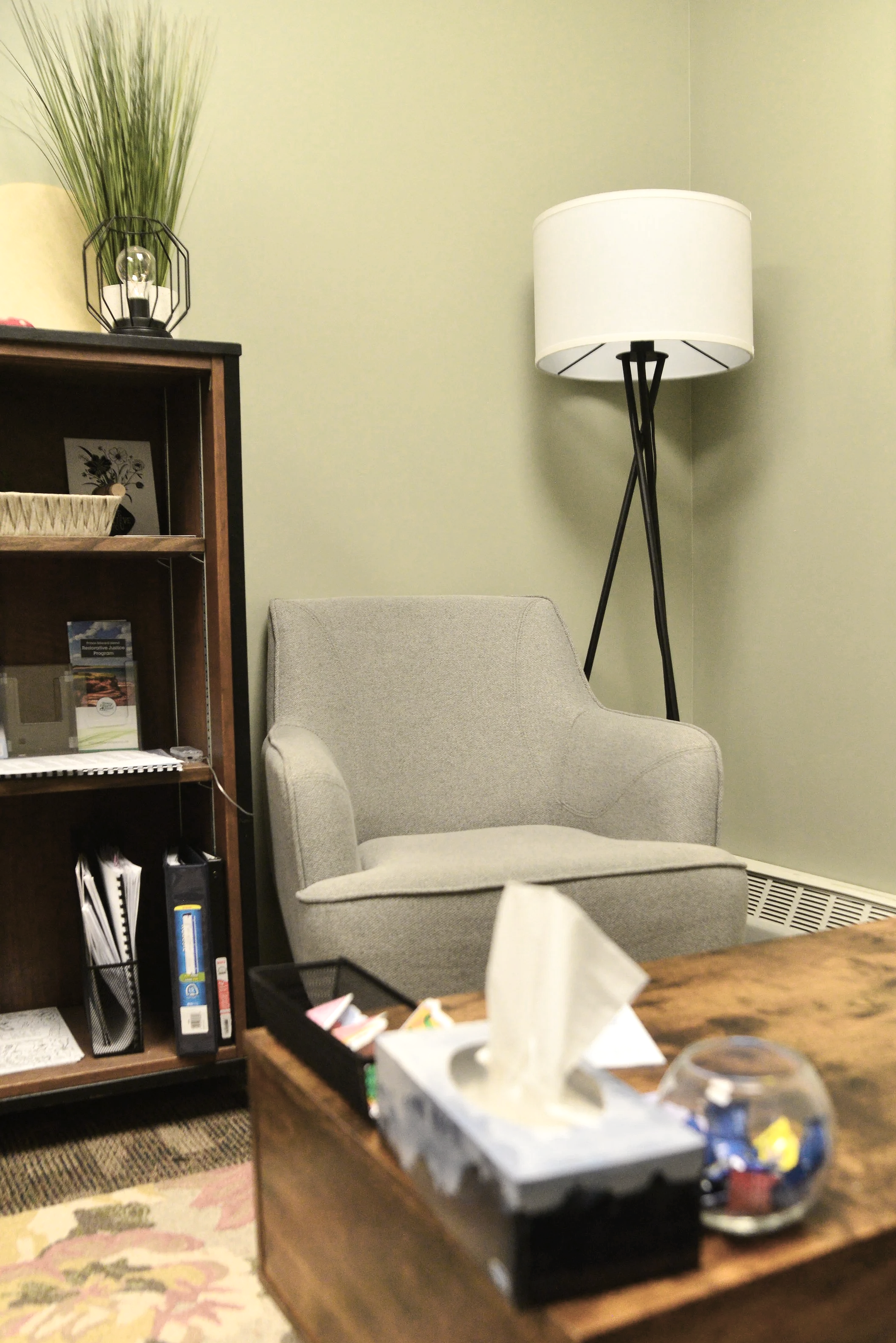 A cozy reading nook with a light gray armchair, a tall modern floor lamp with a white shade, a wooden bookshelf filled with books and decorative items, and a small table with a tissue box and a fishbowl.