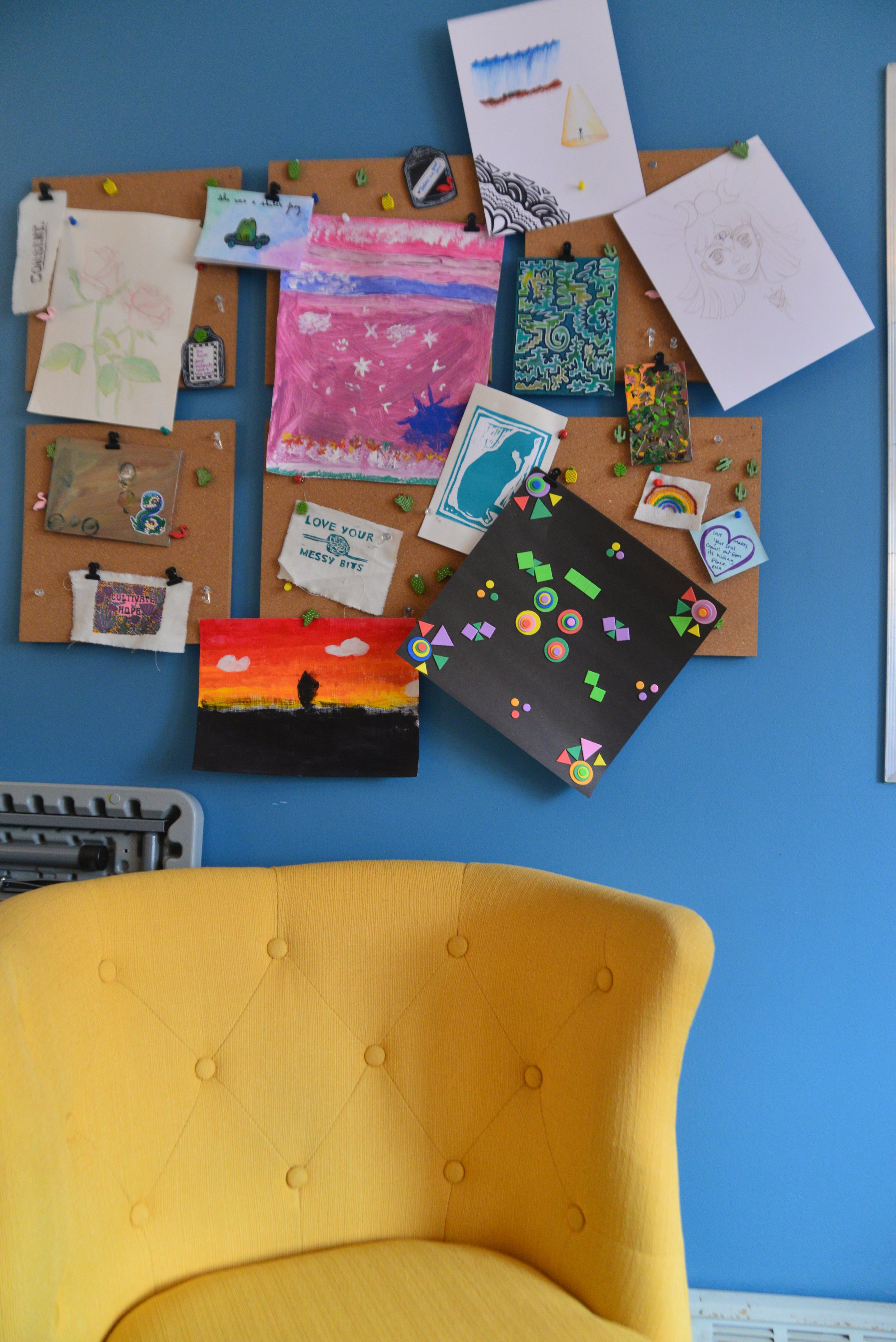Colorful wall of various artwork, drawings, and craft projects pinned to cork boards on a blue wall, with a yellow chair in the foreground.