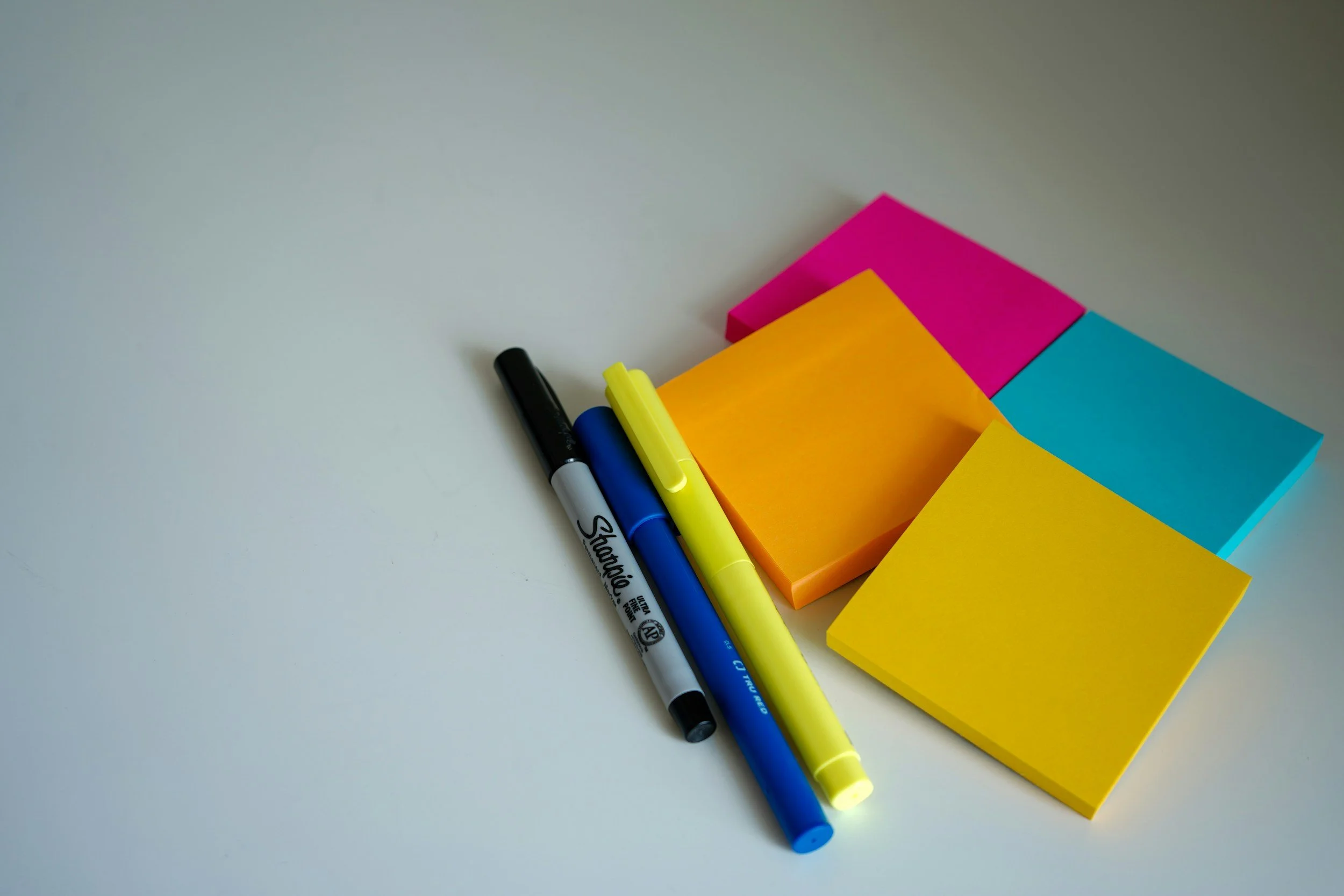 Colorful sticky notes in pink, blue, orange, and yellow with three markers in black, blue, and yellow on a white surface.