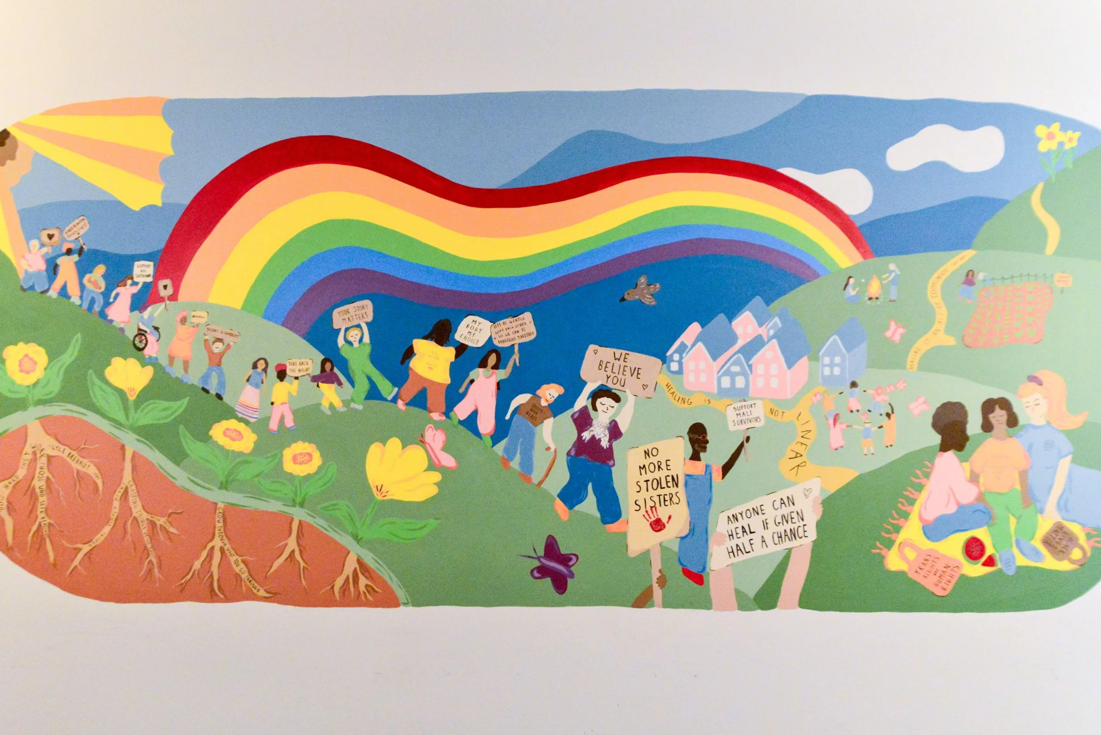 A colorful mural depicting a vibrant community scene with people holding signs advocating for equality and support, a rainbow in the sky, hills with trees and flowers, and various activities symbolizing unity and hope.