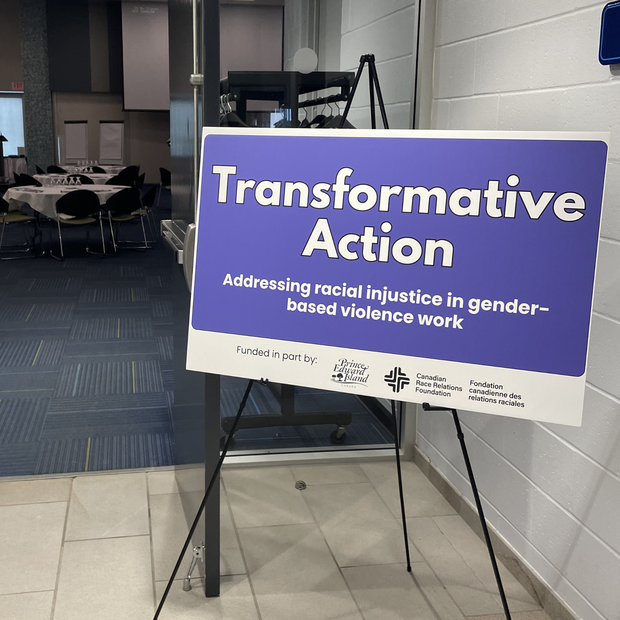 Signboard titled 'Transformative Action' with a subtitle about addressing racial injustice in gender-based violence work, sponsored by Prince Edward Island, Canadian Race Relations Foundation, and Fondation canadienne des relations raciales, displayed in an indoor hall with tables and chairs in the background.