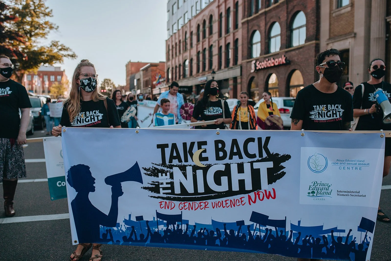 People participating in a protest march holding a large banner reading 'Take Back the Night' and 'End Gender Violence Now' on a city street with storefronts and buildings in the background. Participants are wearing masks.