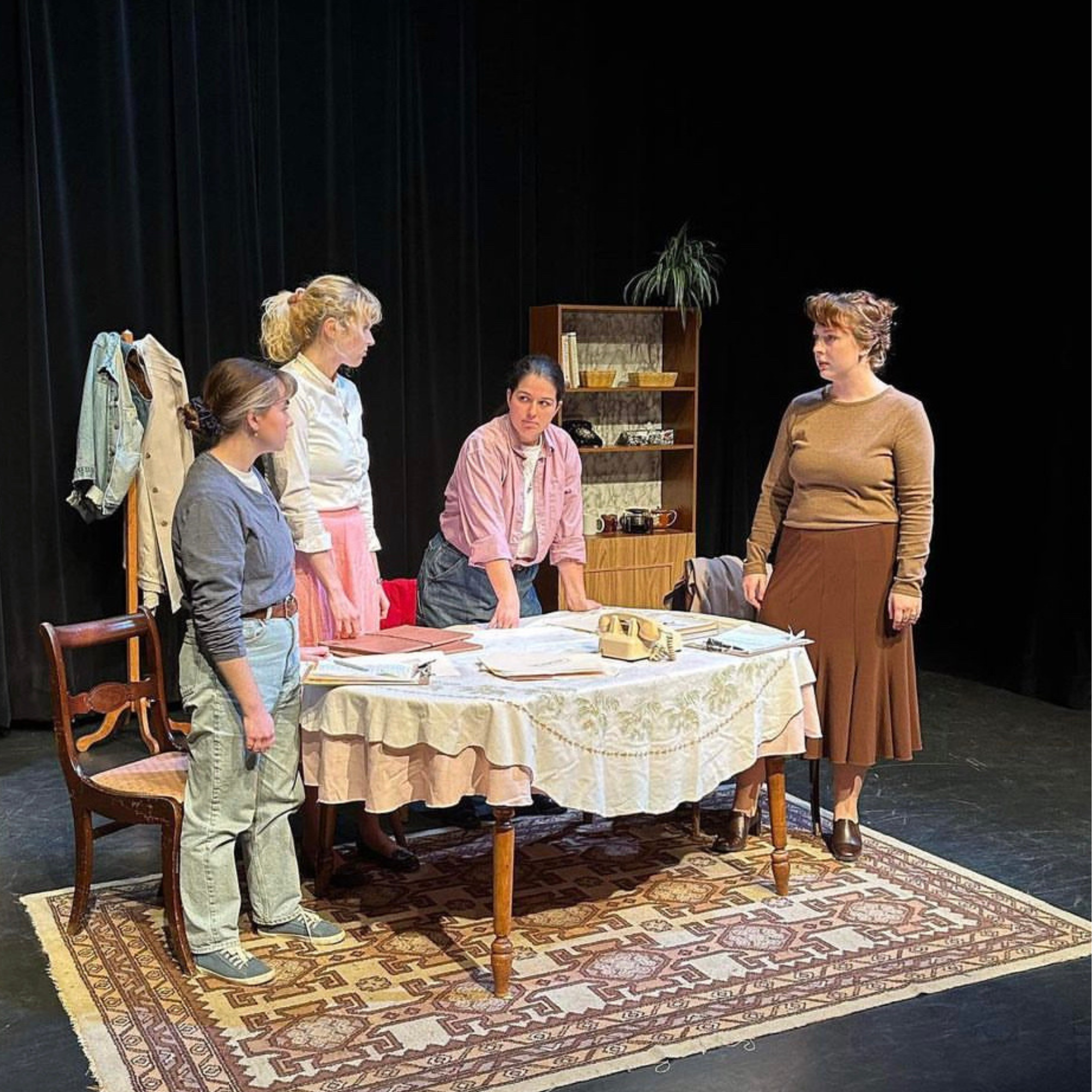 Four women in a room with black curtains, gathered around a table with papers, a rotary phone, and a tablecloth. One woman stands at the end of the table, three women face her, with a coat rack and bookshelf in the background.