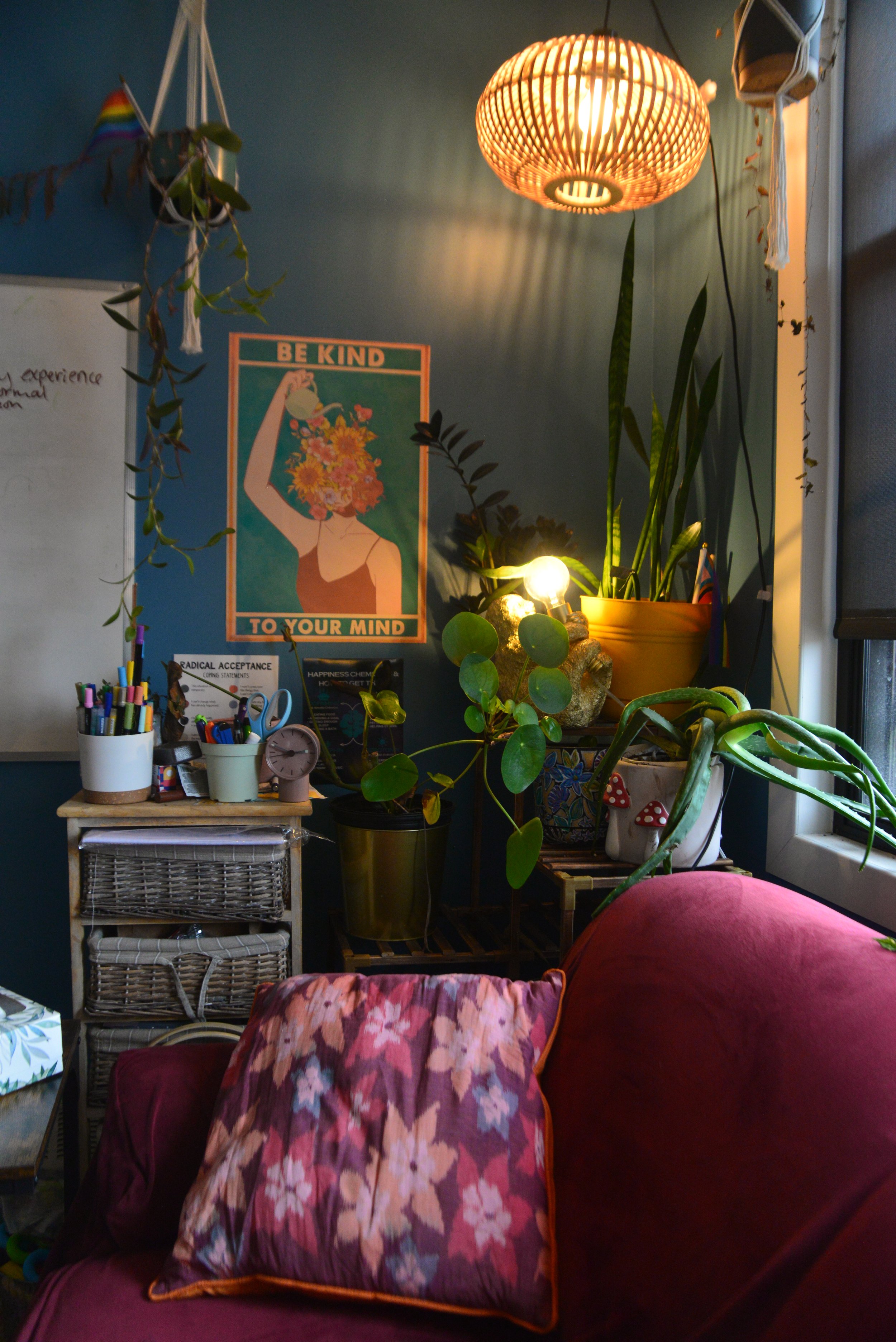 A cozy indoor space with a deep pink velvet sofa and floral pillow. Behind the sofa, a wall with blue paint, decorated with a colorful poster that reads 'Be Kind to Your Mind.' The space features several potted plants, a wicker shelving unit, and a table holding various stationery supplies. A warm, woven pendant light hangs from the ceiling, illuminating the corner.