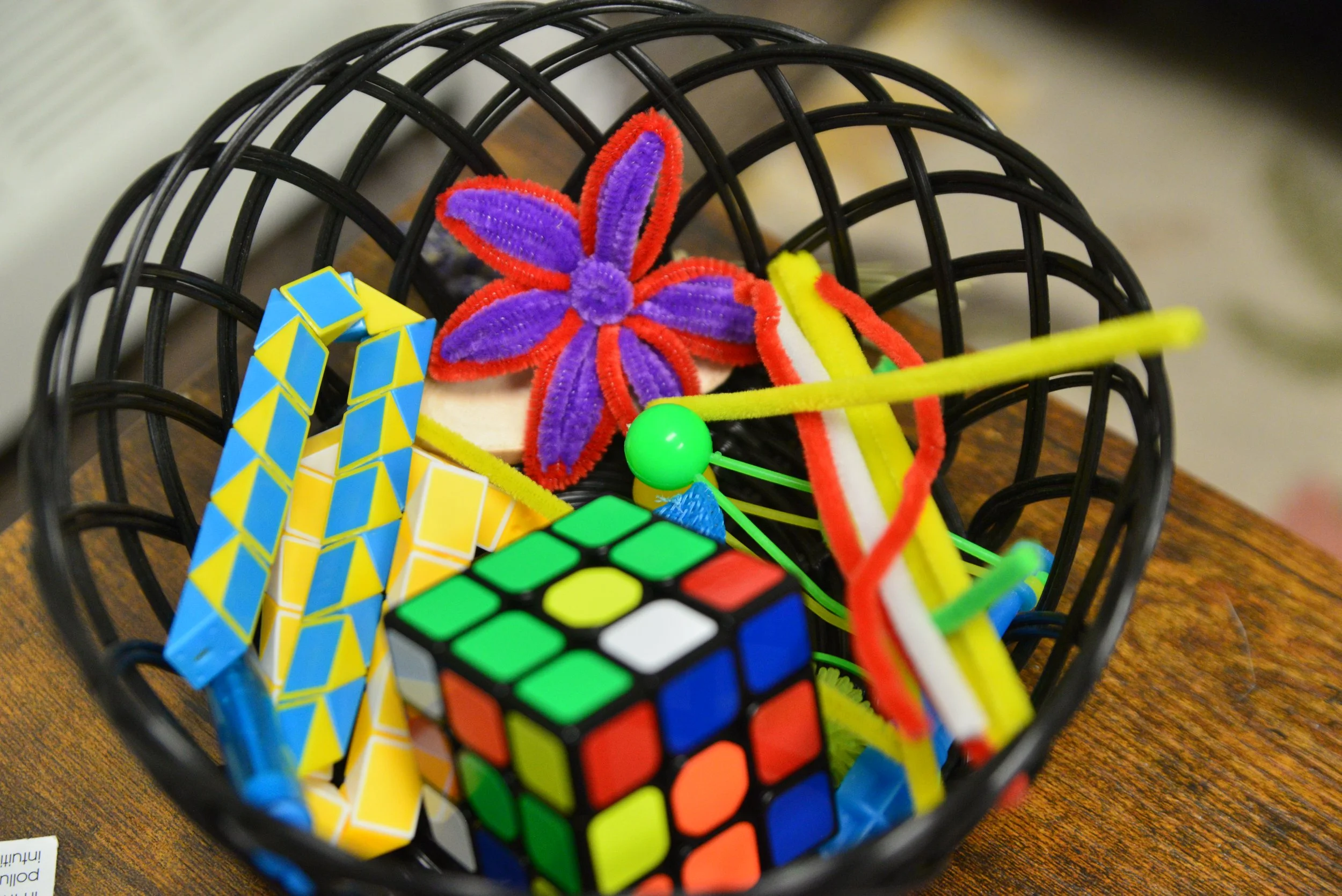 A black wire basket containing colorful children's toys including a Rubik's cube, pipe cleaners, a foam flower, and a stegosaurus-shaped puzzle piece.