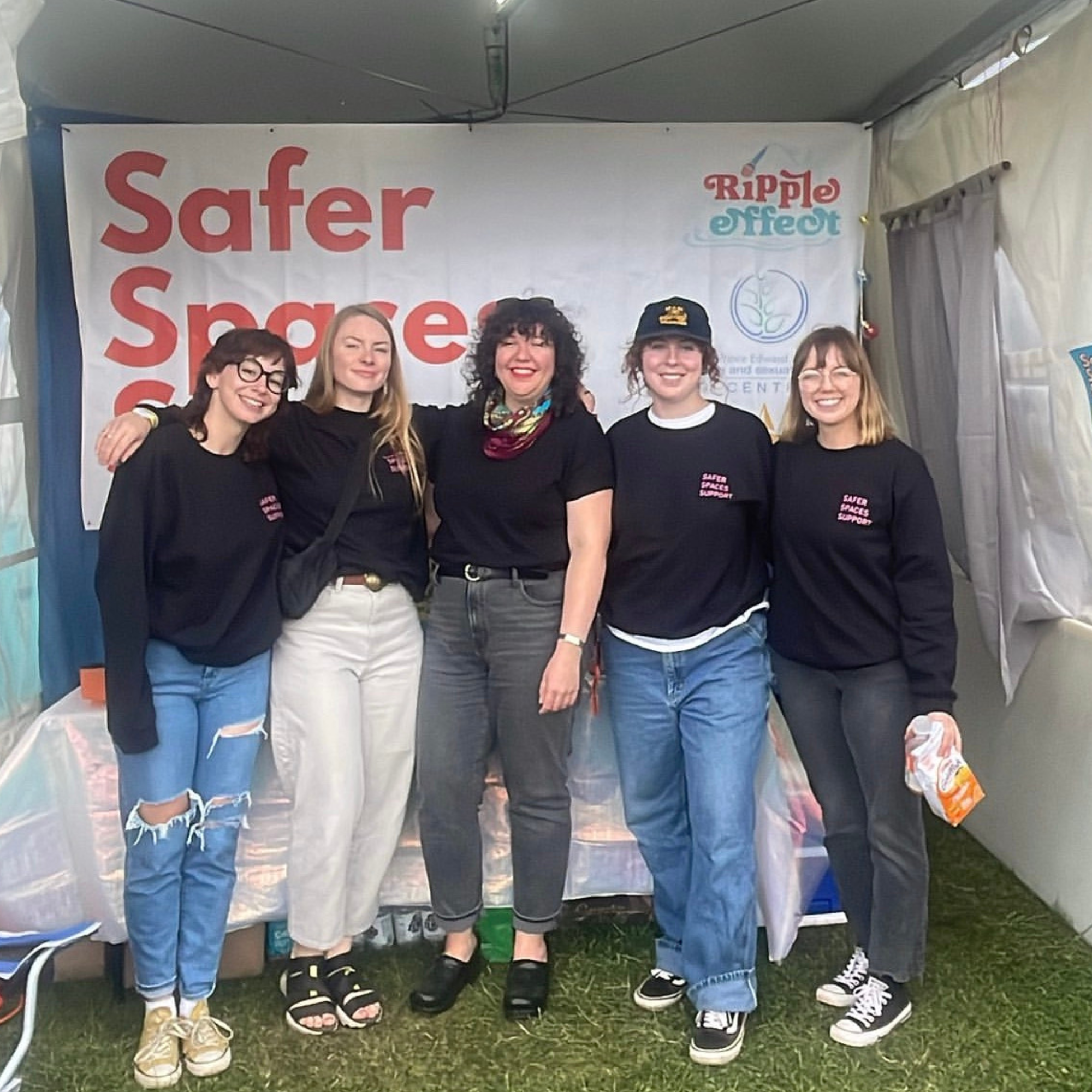 Group of five women standing together at an event, smiling, in front of a banner that reads 'Safer Space' and 'Ripple Effect.'