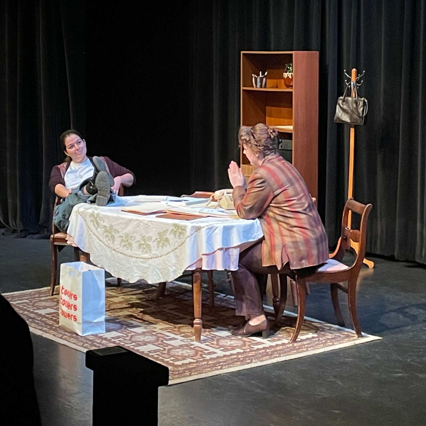 Two women sitting at a dining table with papers and a telephone, engaged in conversation in a room with black curtains and a wooden shelf.