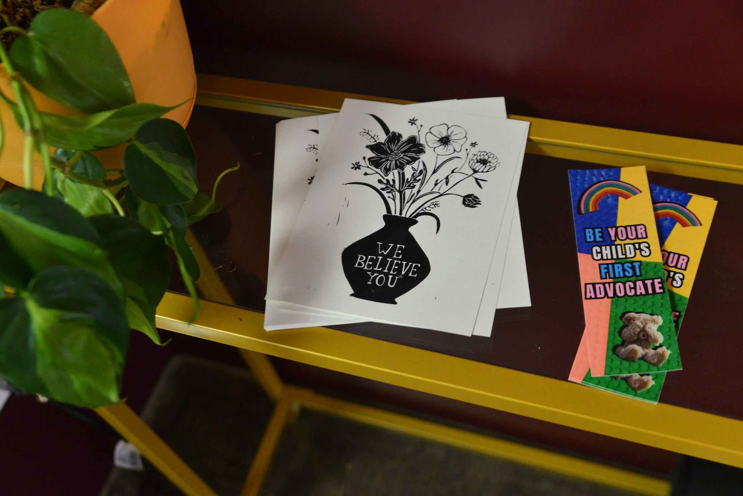 Black-and-white coloring page featuring a vase with flowers and the words "We Believe You." Colorful advocacy stickers with rainbows and teddy bears, themes of support and advocacy, are also visible.