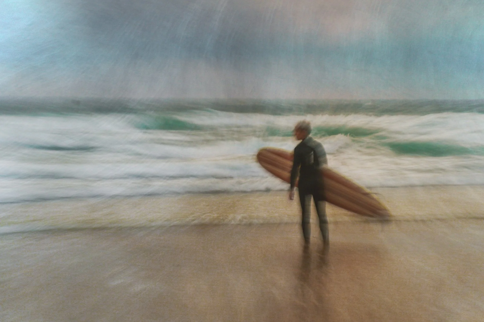 Image of a person in a wetsuit holding a surfboard on a beach with ocean waves in the background.