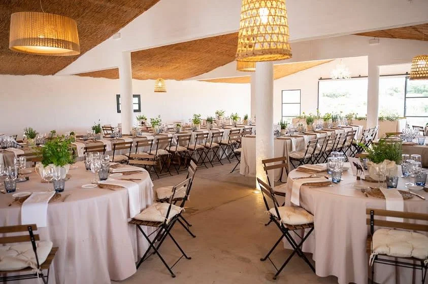 Salón de evento decorado con mesas redondas con manteles blancos y centros de flores verdes, sillas de madera con cojines, iluminación cálida y ventanas grandes que dejan entrar luz natural.