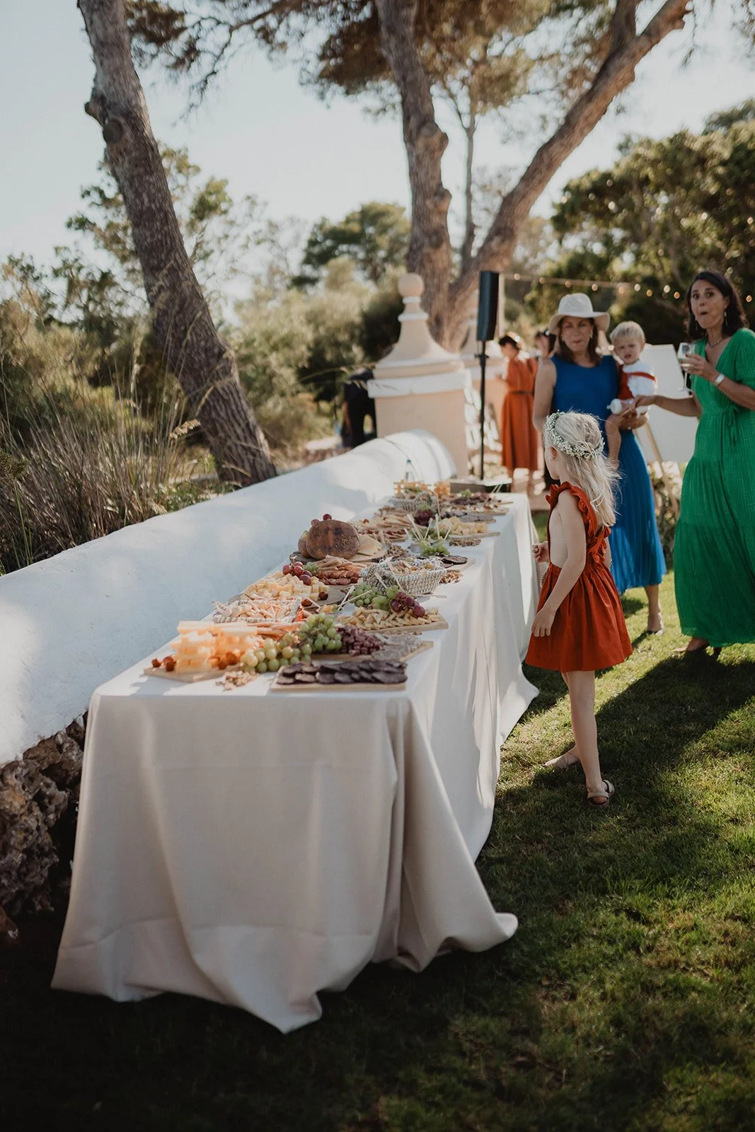 Mesa de aperitivos en una fiesta al aire libre, con niños y adultos en el fondo, en un entorno natural con árboles y césped.