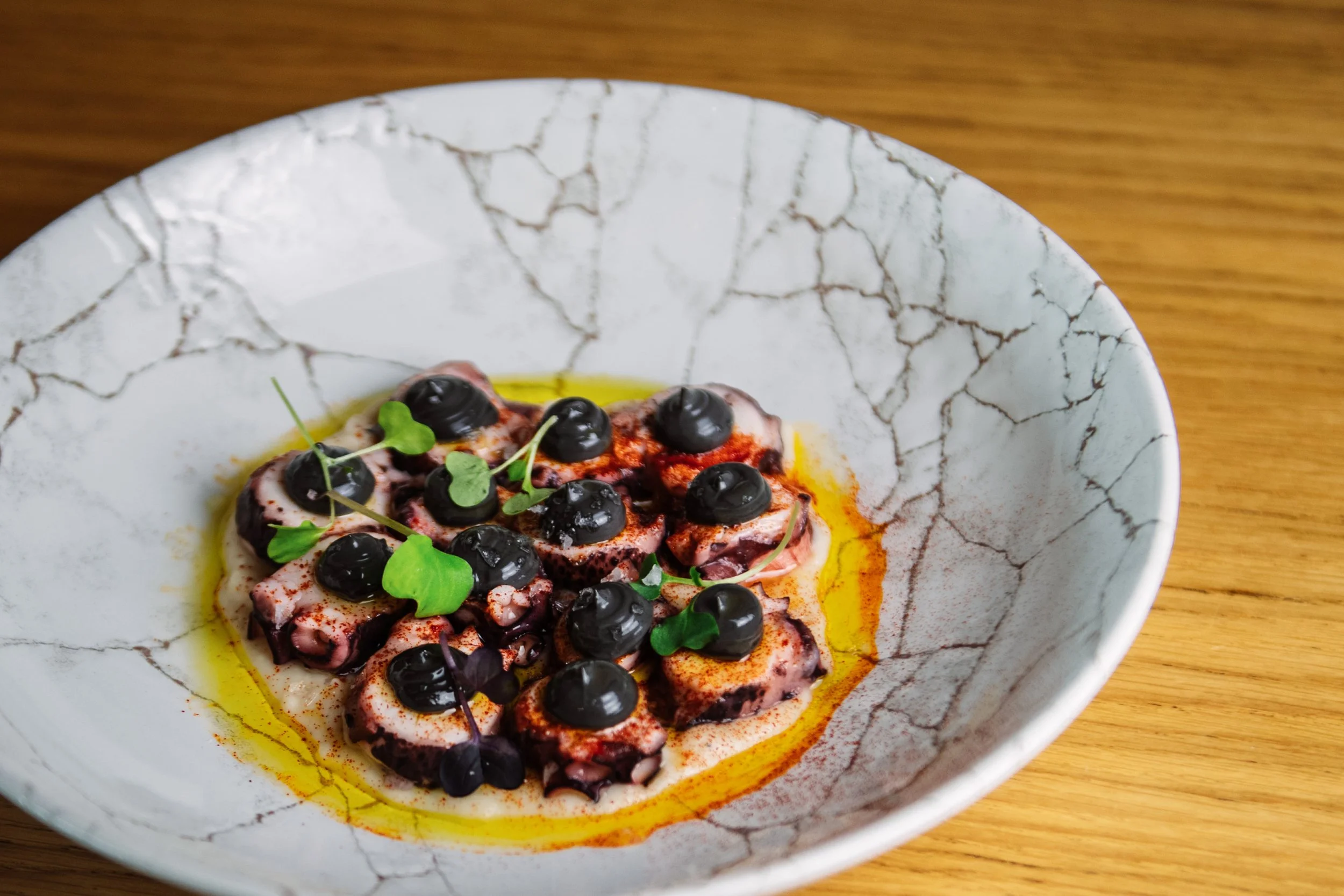 Plato de pulpo con aceitunas negras, aceite de oliva y hierbas frescas en un cuenco de cerámica con grietas decorativas.
