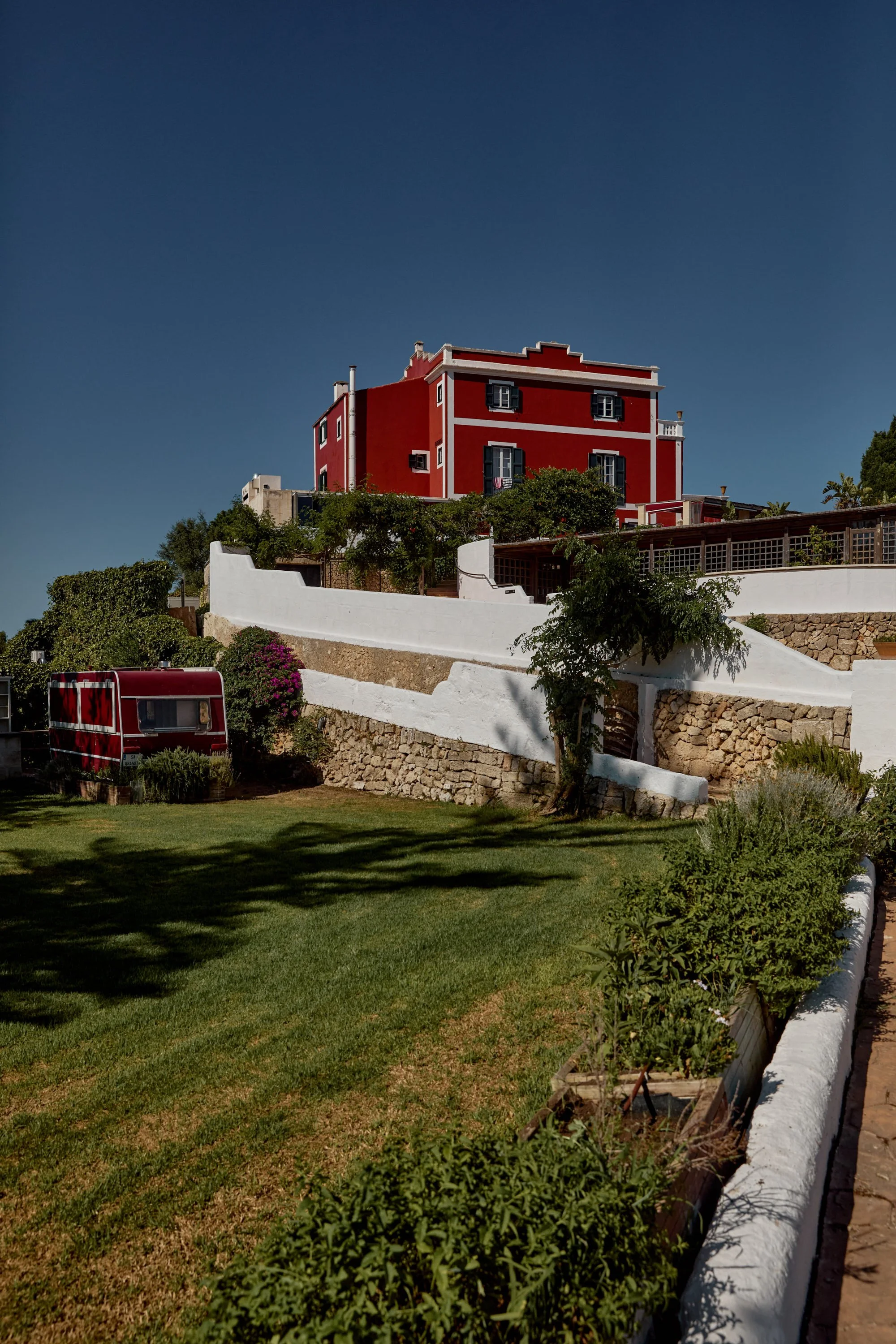 Casa color rojo en una colina, con muros blancos y vegetación, bajo un cielo despejado.