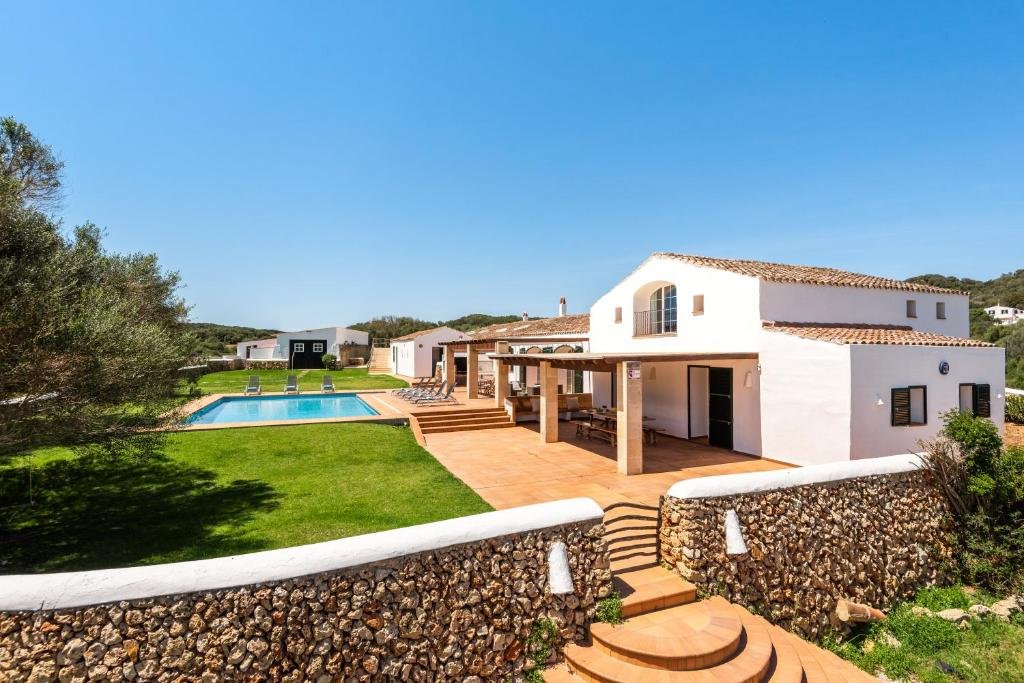 Casa blanca con techo de tejas, patio con pérgola, piscina y áreas verdes, en un entorno tranquilo y soleado.