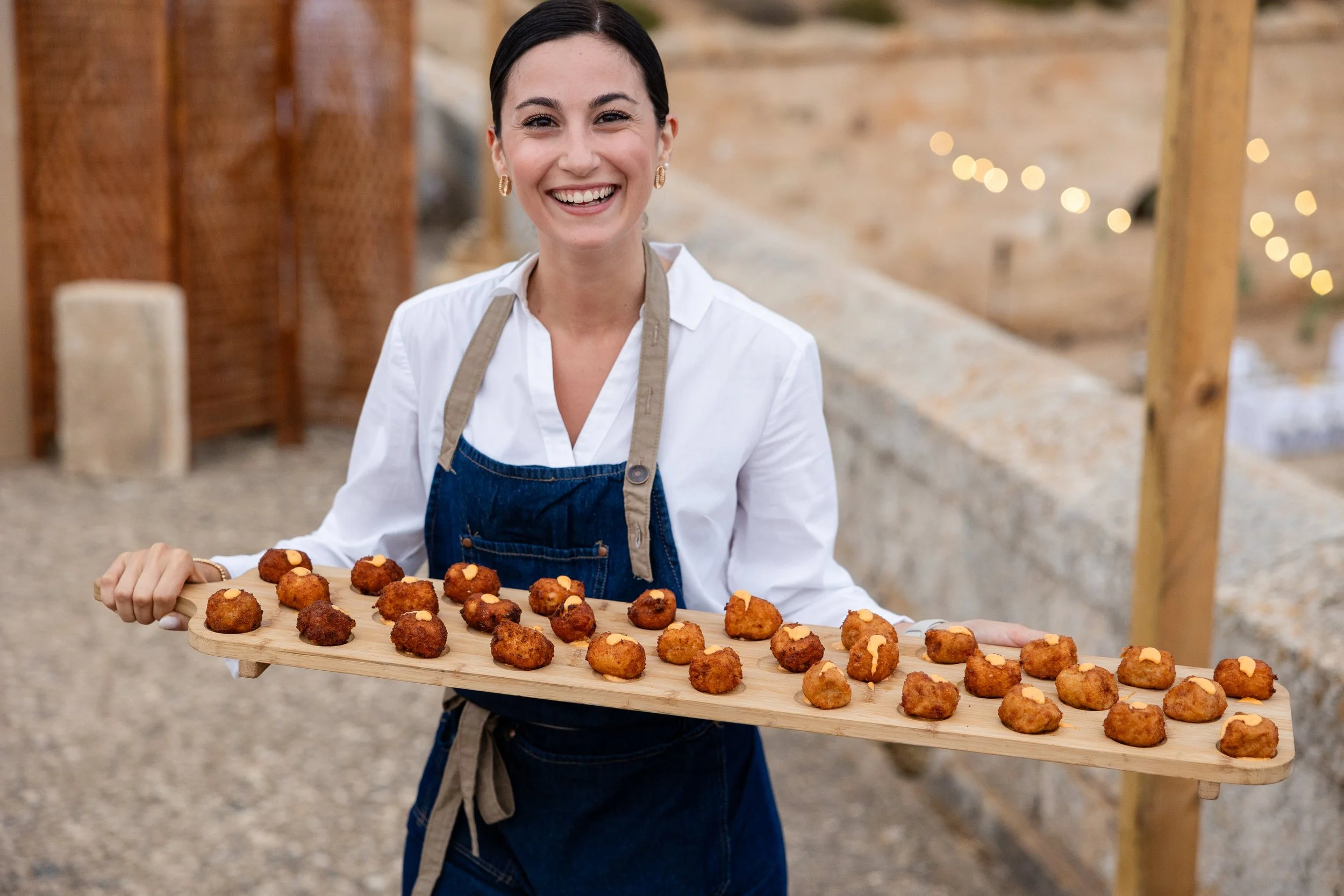 Mujer sonriente sosteniendo una bandeja con croquetas de pollo en un ambiente al aire libre, decorado con luces pequeñas.