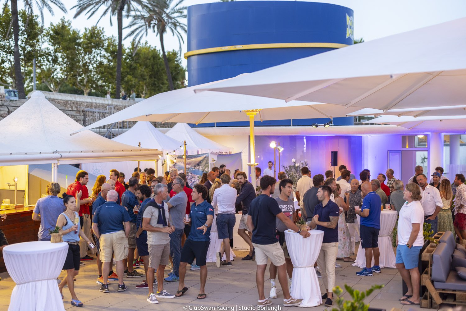 Reunión social al aire libre con muchas personas, algunas conversando y otras disfrutando bebidas, en un evento en la tarde con toldos y decoraciones luminosas.