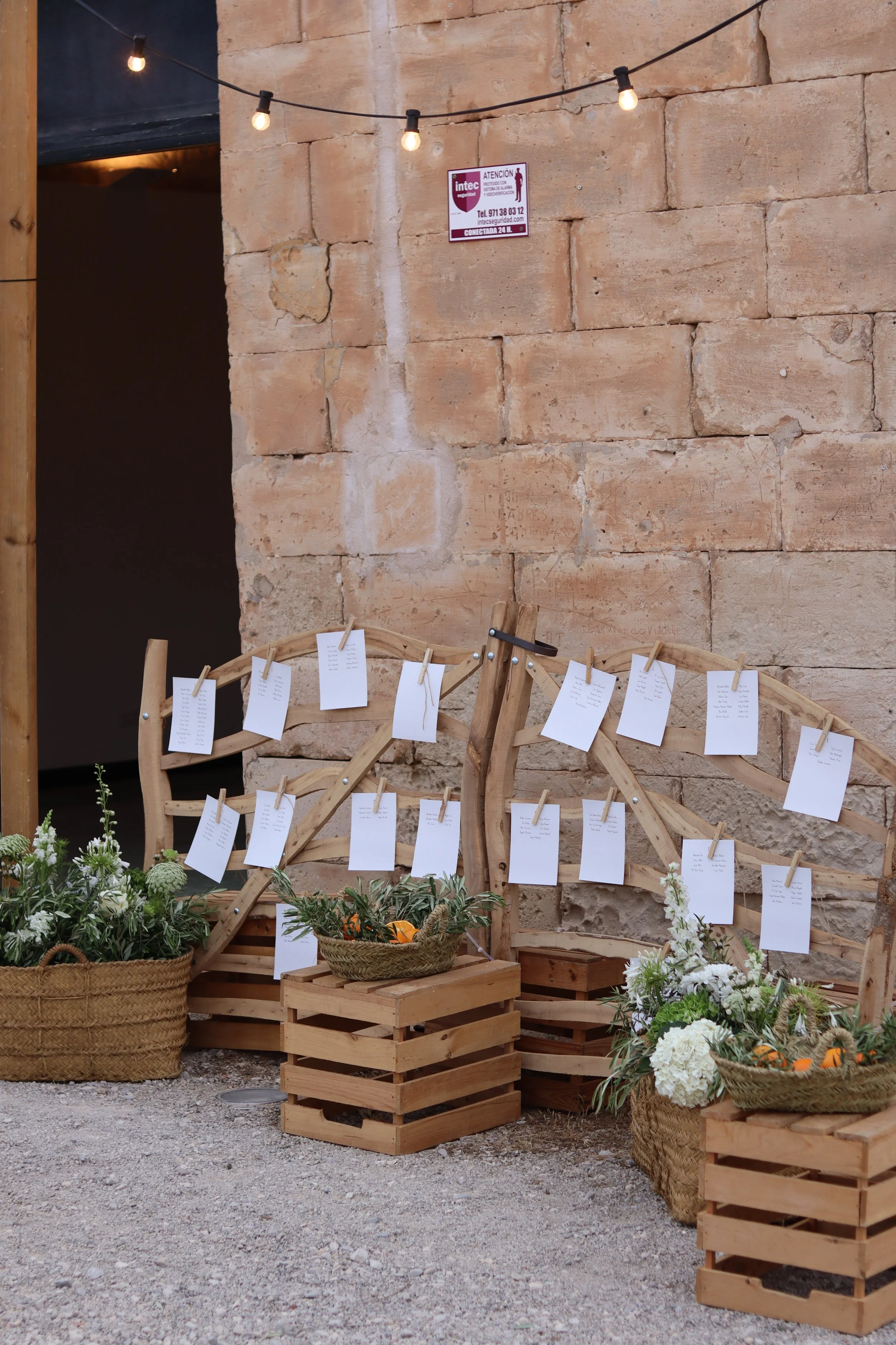 Escenario decorativo con cestas de mimbre y cajas de madera, adornadas con plantas y flores blancas, y con notas de papel colgadas con pinzas, en un ambiente rústico.