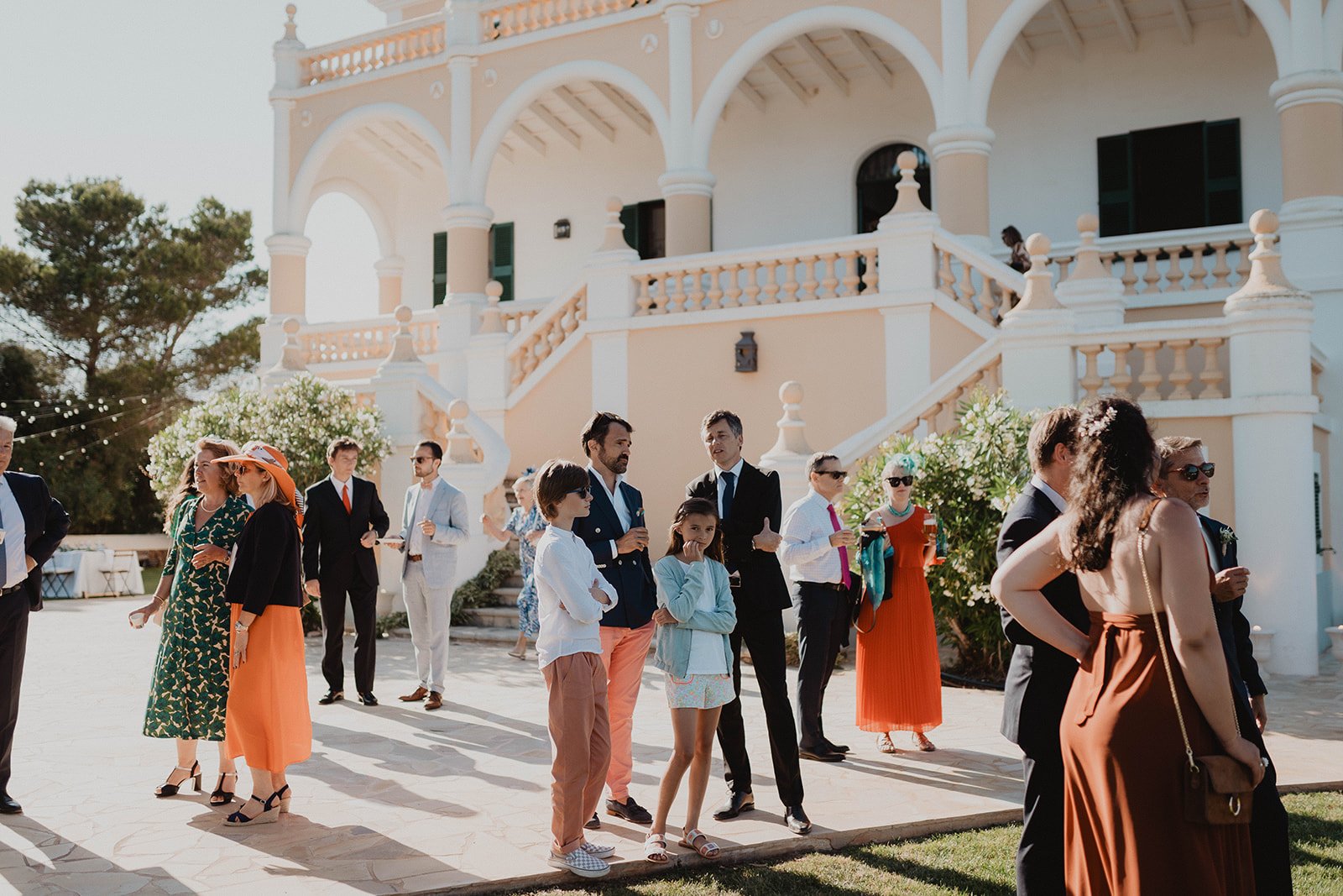 Personas en una fiesta al aire libre frente a un edificio de estilo clásico, con personas conversando, algunos con trajes formales y otros con ropa casual, en un ambiente soleado.
