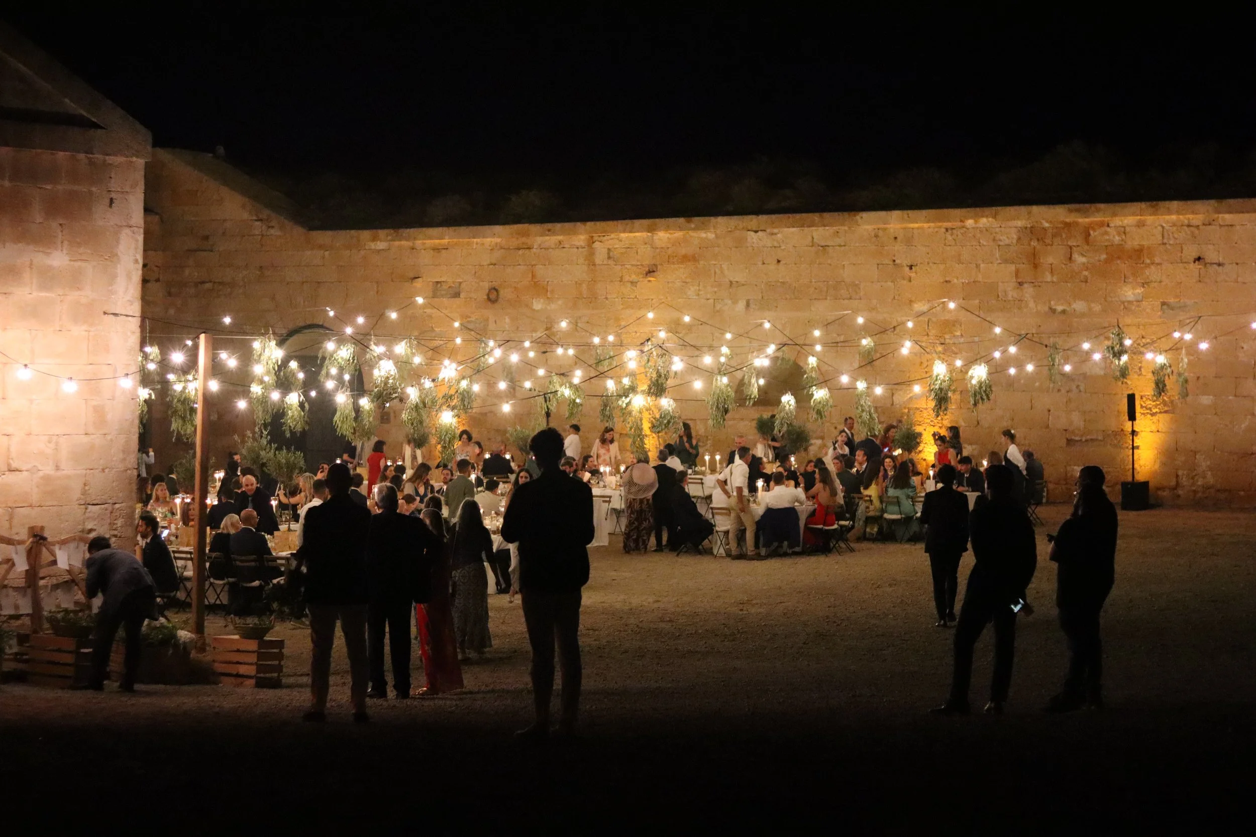 Celebración nocturna con mesas, luces y gente en un evento en un patio con muros de piedra.