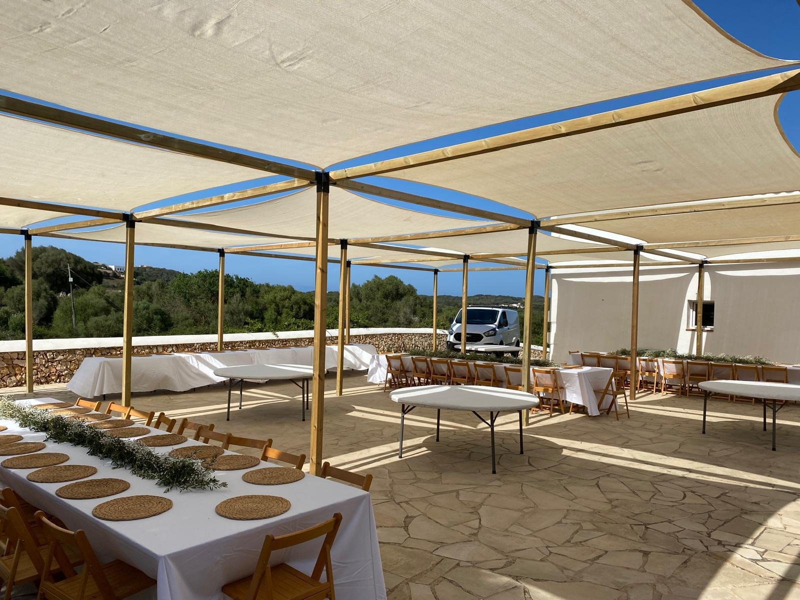 Área al aire libre preparada para un evento, con mesas cubiertas con manteles blancos y sillas de madera, bajo una estructura de lona o tela para sombra, con vegetación y un auto estacionado en el fondo