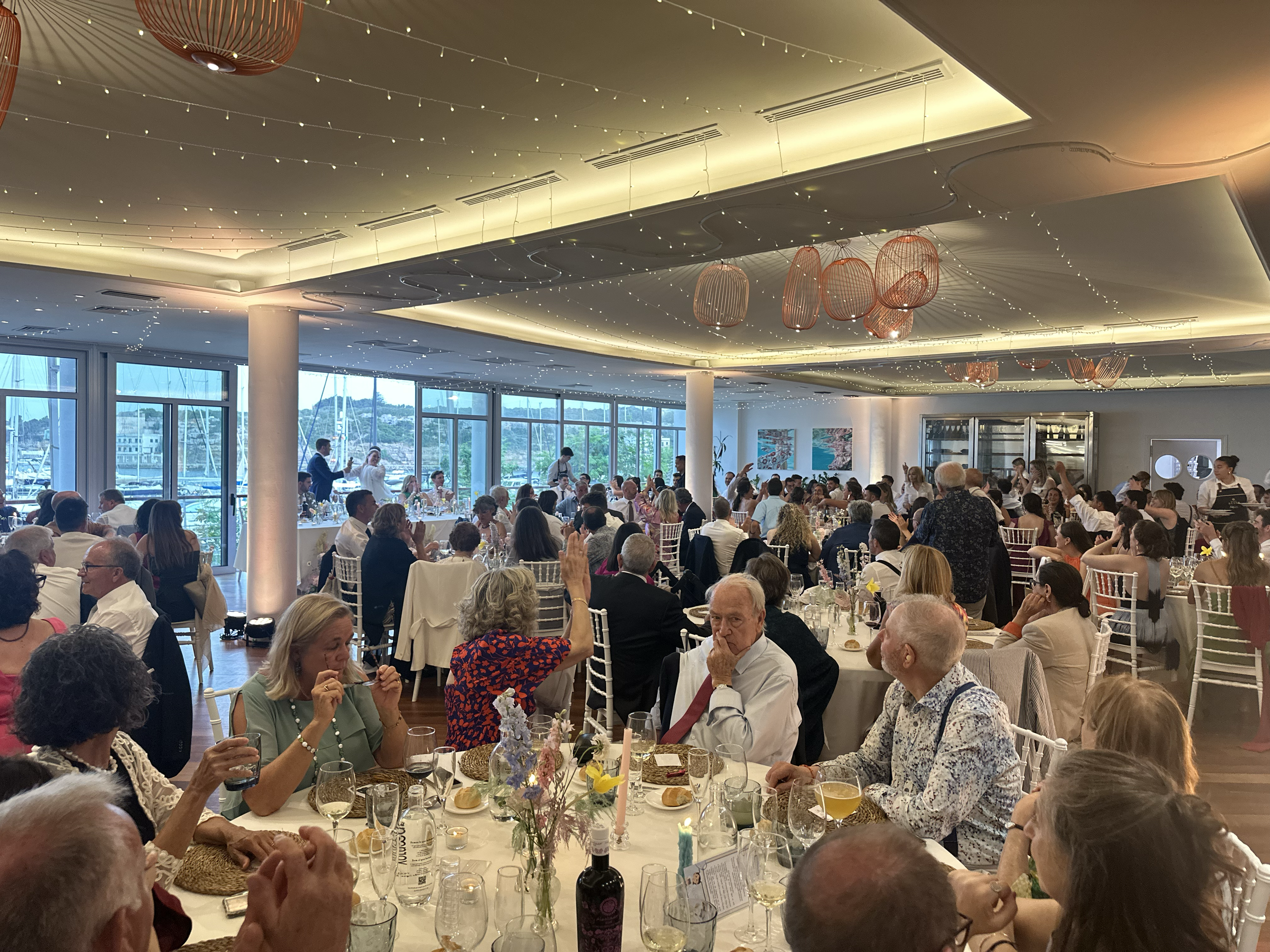 Cena en un restaurante elegante con muchas mesas llenas de comensales, decorada con luces y jarrones flotantes en el techo, con vista al mar y ventanas grandes.