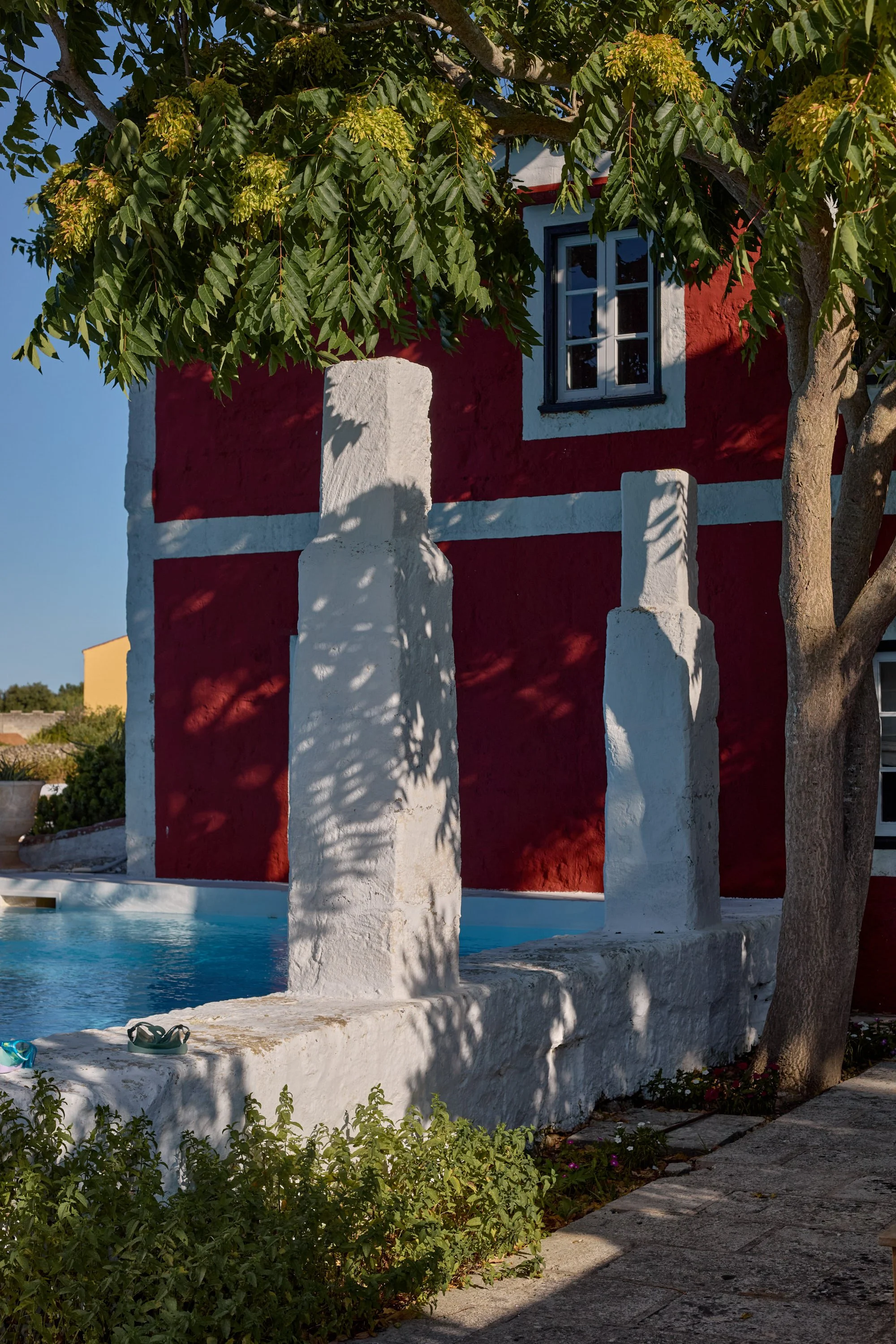 Casa colorida con paredes rojas y ventanas azules, árbol con hojas verdes, fuente de piedra, y piscina, en un entorno soleado.