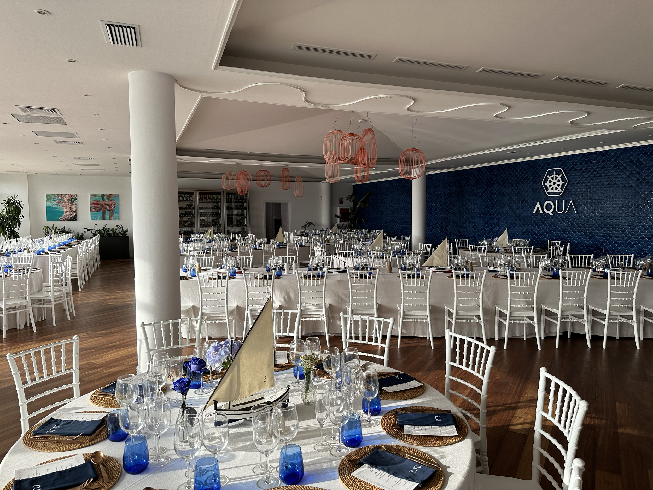 Salón de eventos decorado con mesas blancas, sillas blancas, cristalería y manteles blancos, con un fondo azul y el logo 'Aqua'.