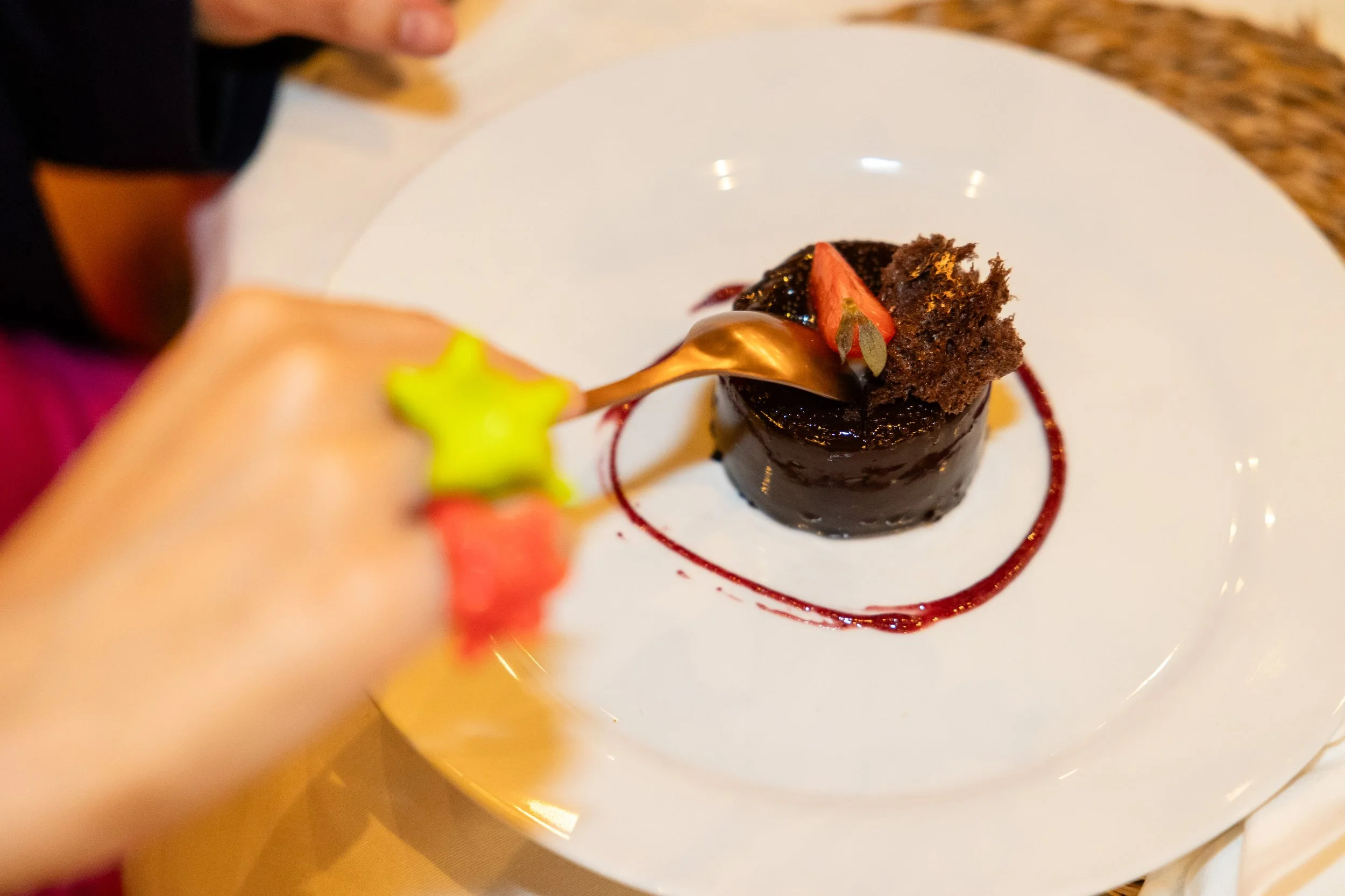 Postre de chocolate con una decoración de fruta y hojas, servido en un plato blanco con salsa de frambuesa y utensilio dorado.
