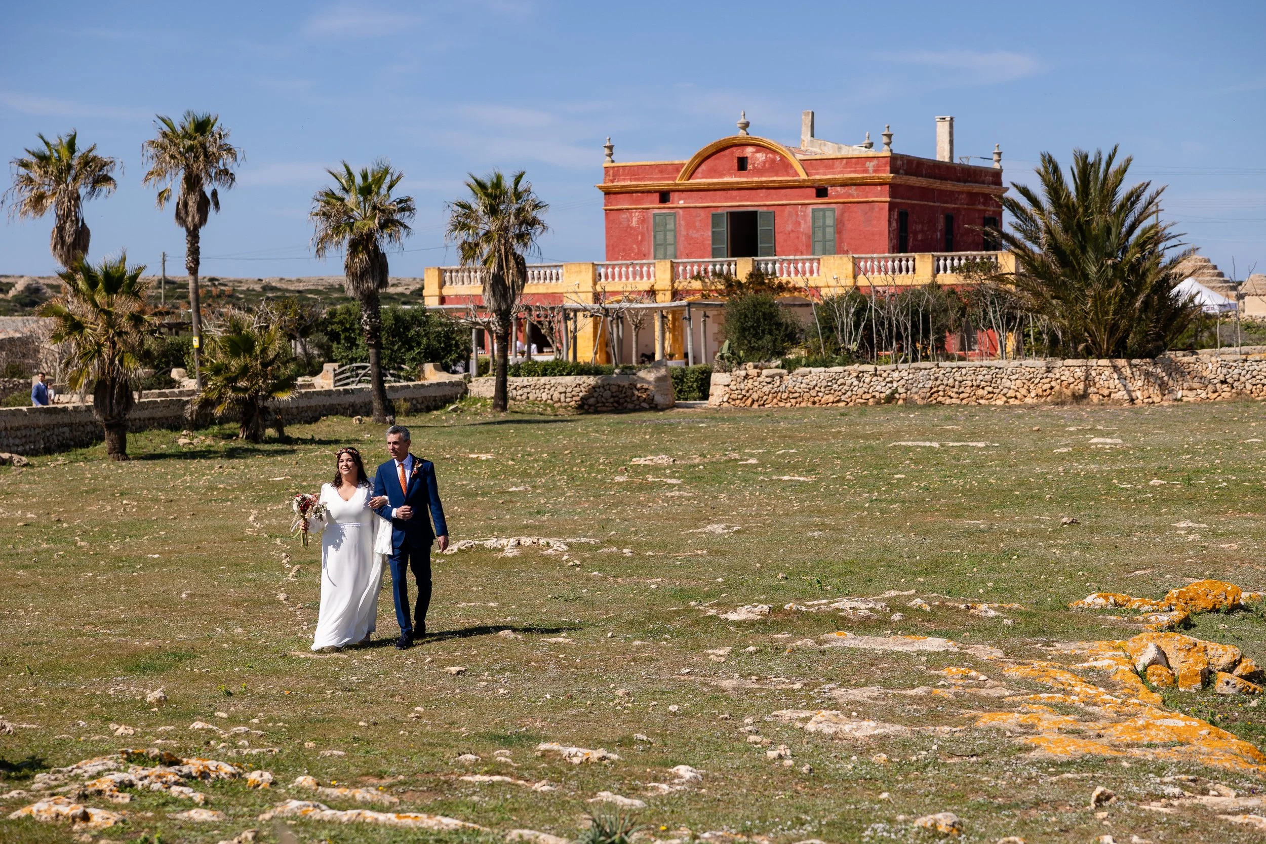 Una pareja de boda caminando en un campo con un edificio color terracota y palmeras en el fondo