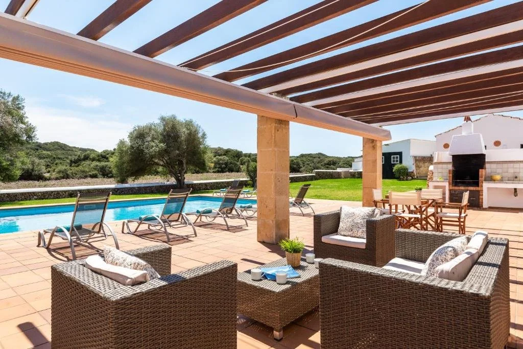 Área de piscina al aire libre con muebles de exterior, sombrillas y zona de barbacoa en un entorno rural con árboles y casas blancas.