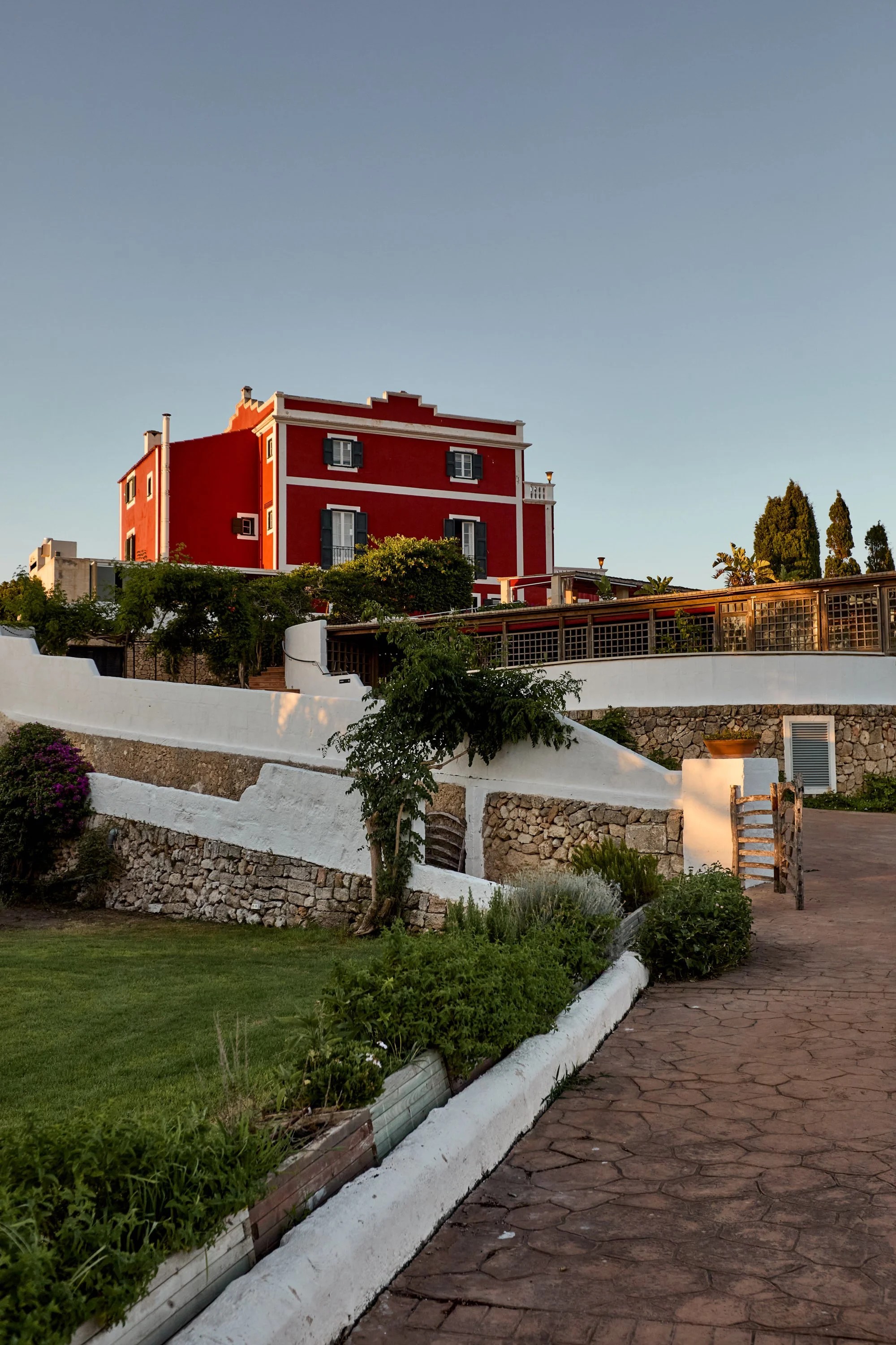 Edificio de color rojo con ventanas y balcones, rodeado de vegetación y muro de piedra, bajo un cielo despejado.