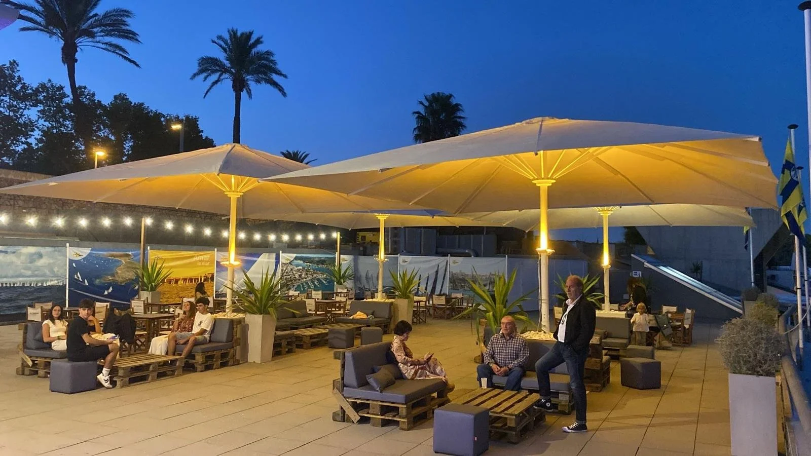 Área de descanso al aire libre en un restaurante o cafetería, con sombrillas grandes, mesas y sillas tipo palets, plantas en macetas, varias personas sentadas conversando y una persona de pie, en la noche con iluminación ambiente y un fondo de mural con imágenes marinas y palmeras.
