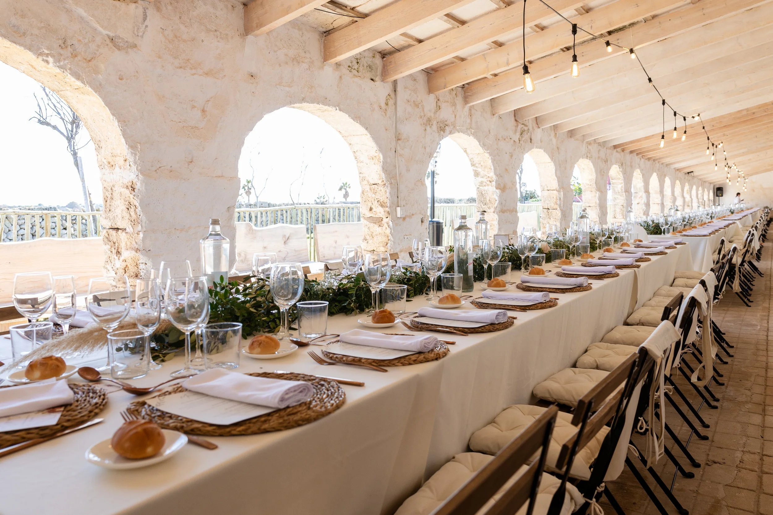 Mesa larga decorada para una celebración, con platos, copas de vino, pan, cubiertos, y centros de plantas verdes, en un lugar con paredes de piedra y arcos, con ventanas abiertas y vista al exterior.