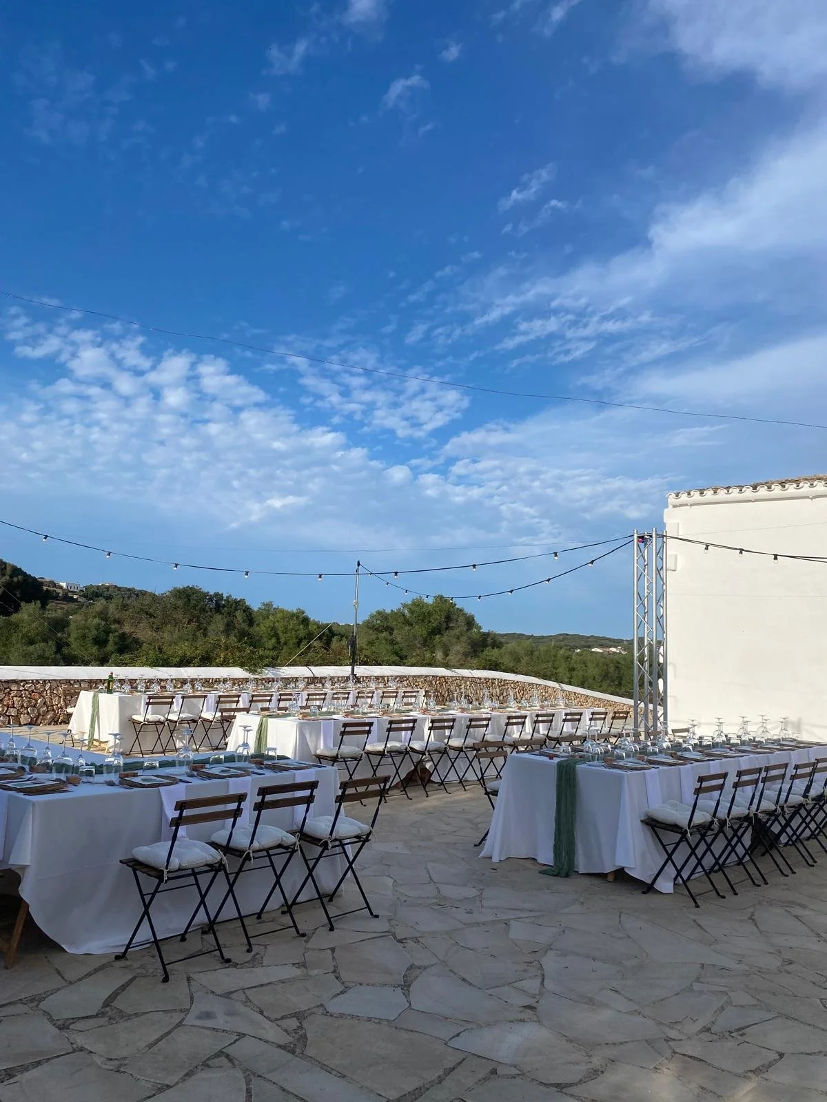 Mesas preparadas para un evento al aire libre con sillas, cristalería y cubiertos, bajo un cielo azul con nubes y decoraciones de luces.