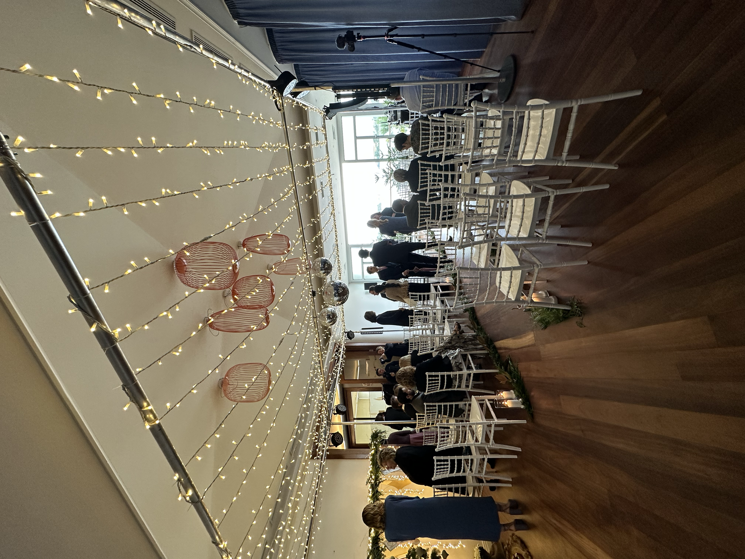 Gente en una fiesta de celebración con mesas y sillas blancas en un salón decorado con cadenas de luces y adornos navideños.