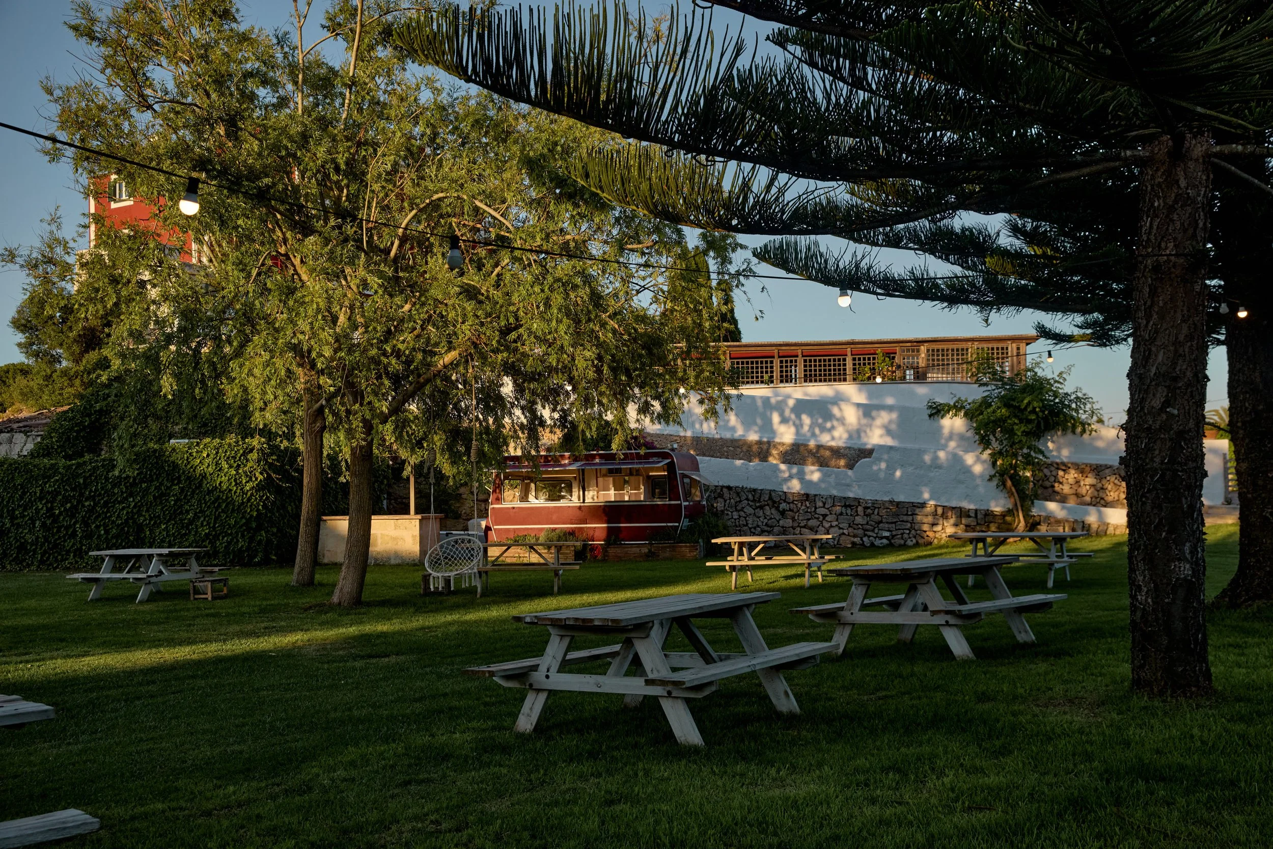 Jardín con mesas de picnic de madera, árboles y una germinadora roja, en la tarde con luz suave y cadenas de luces colgadas.