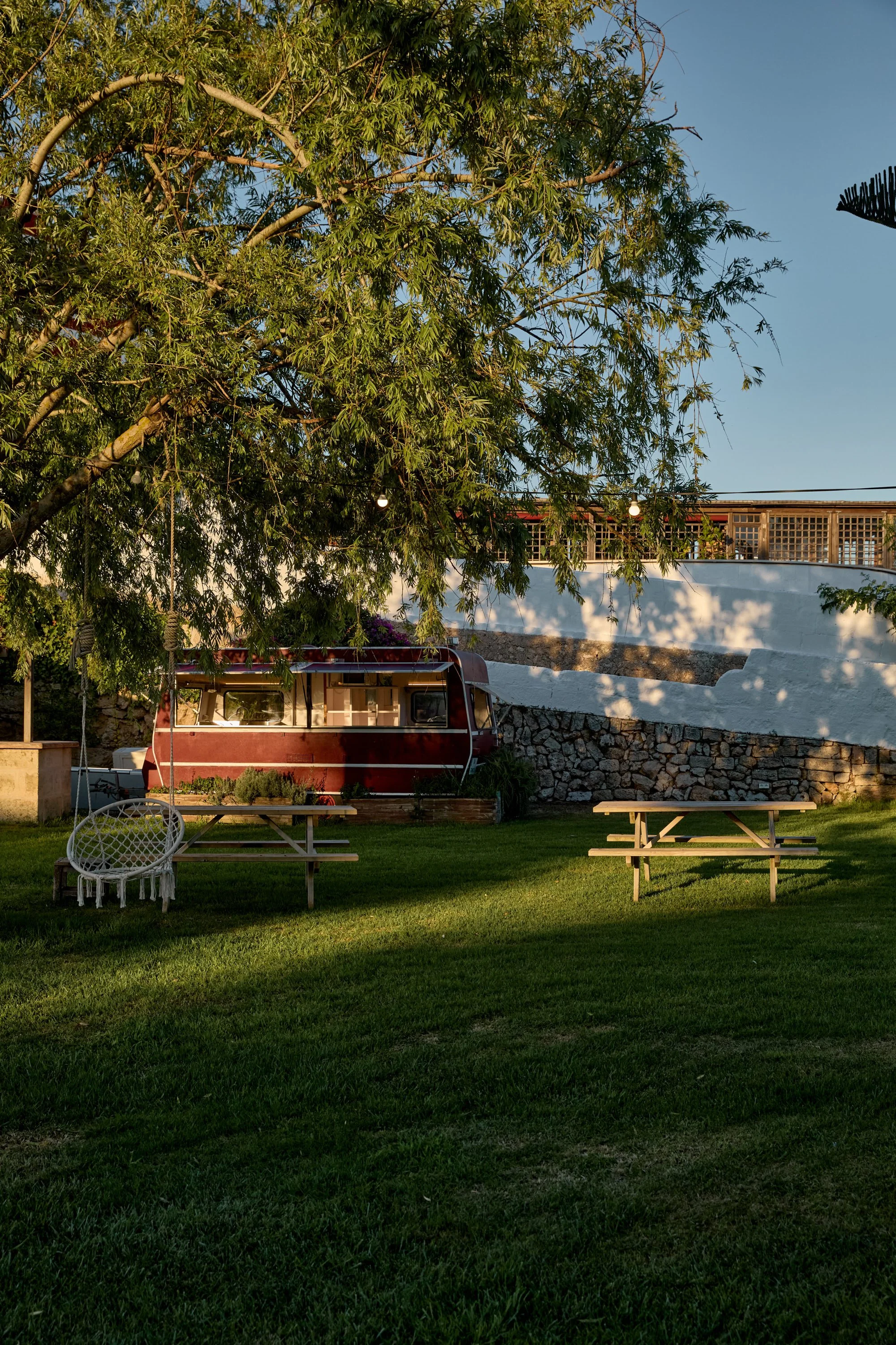 Un jardín con césped verde, un árbol grande con ramas frondosas, dos mesas de picnic de madera, una silla colgante de mimbre y un remolque pintado de rojo estacionado frente a una cerca y una pared blanca con piedra en la base, en un día soleado con 