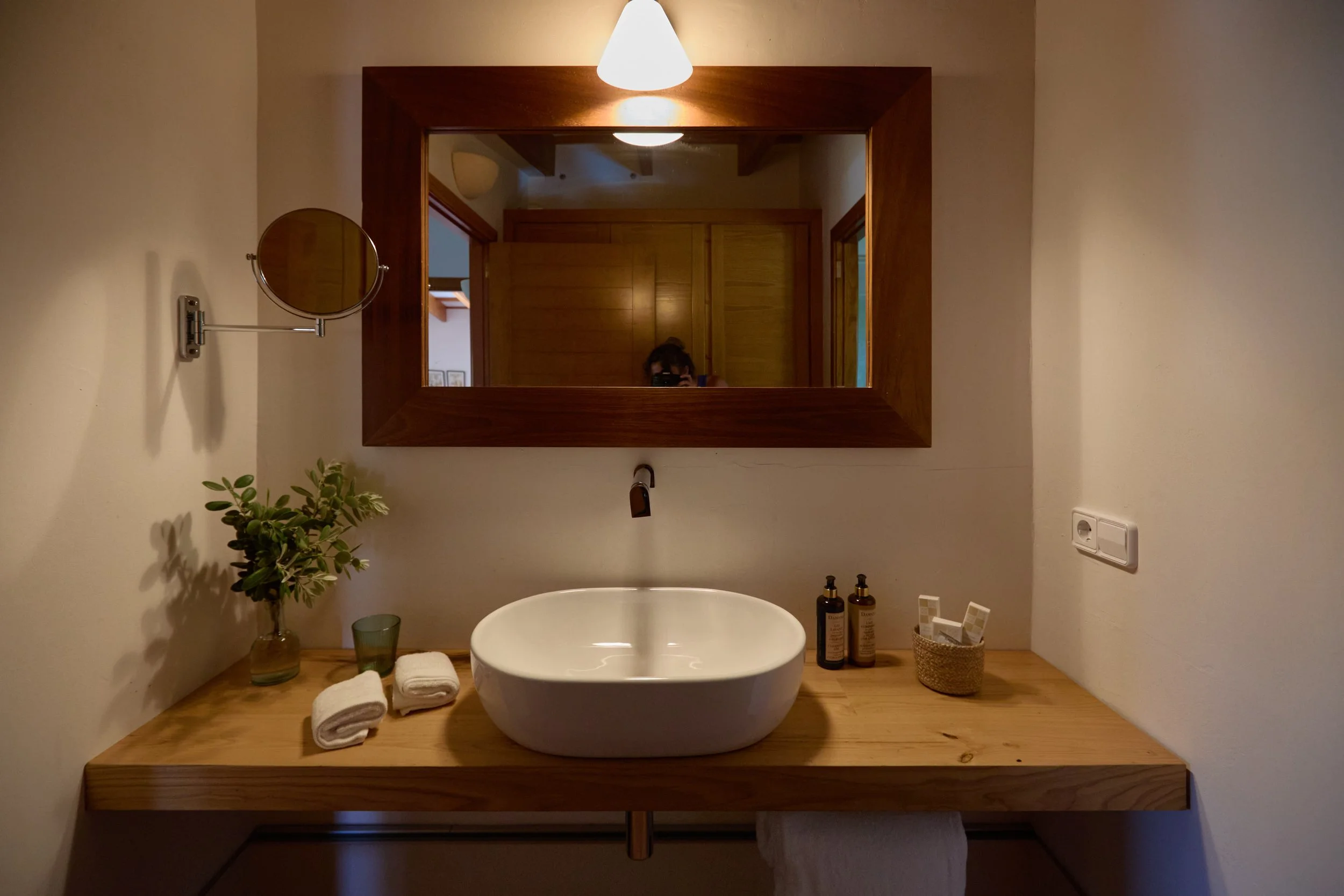  lavabo de cerámica blanco sobre una encimera de madera, con un espejo grande de marco de madera, una planta en un vaso, toallas enrolladas, y productos de aseo, en un baño con paredes claras y luz cálida. 
