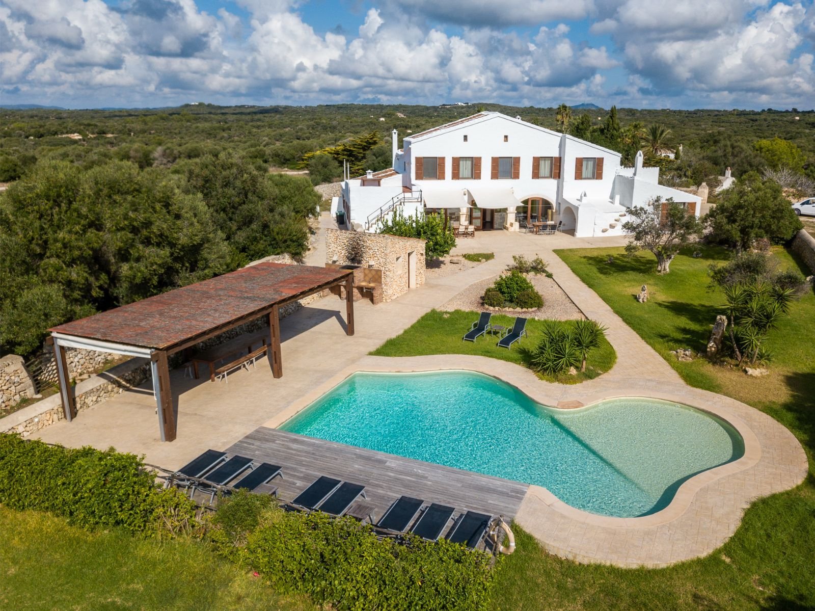 Vista aérea de una casa blanca con techos rojos, piscina en forma de corazón, área de descanso con sombrillas, césped, árboles y un camino de piedra en un entorno natural.