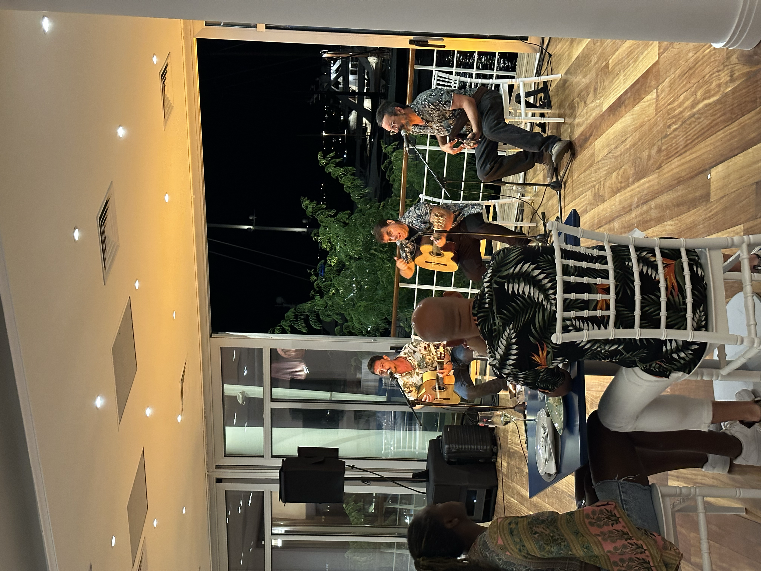 Un grupo de músicos tocando guitarras en una presentación al aire libre por la noche, con un público sentado en sillas blancas observando.