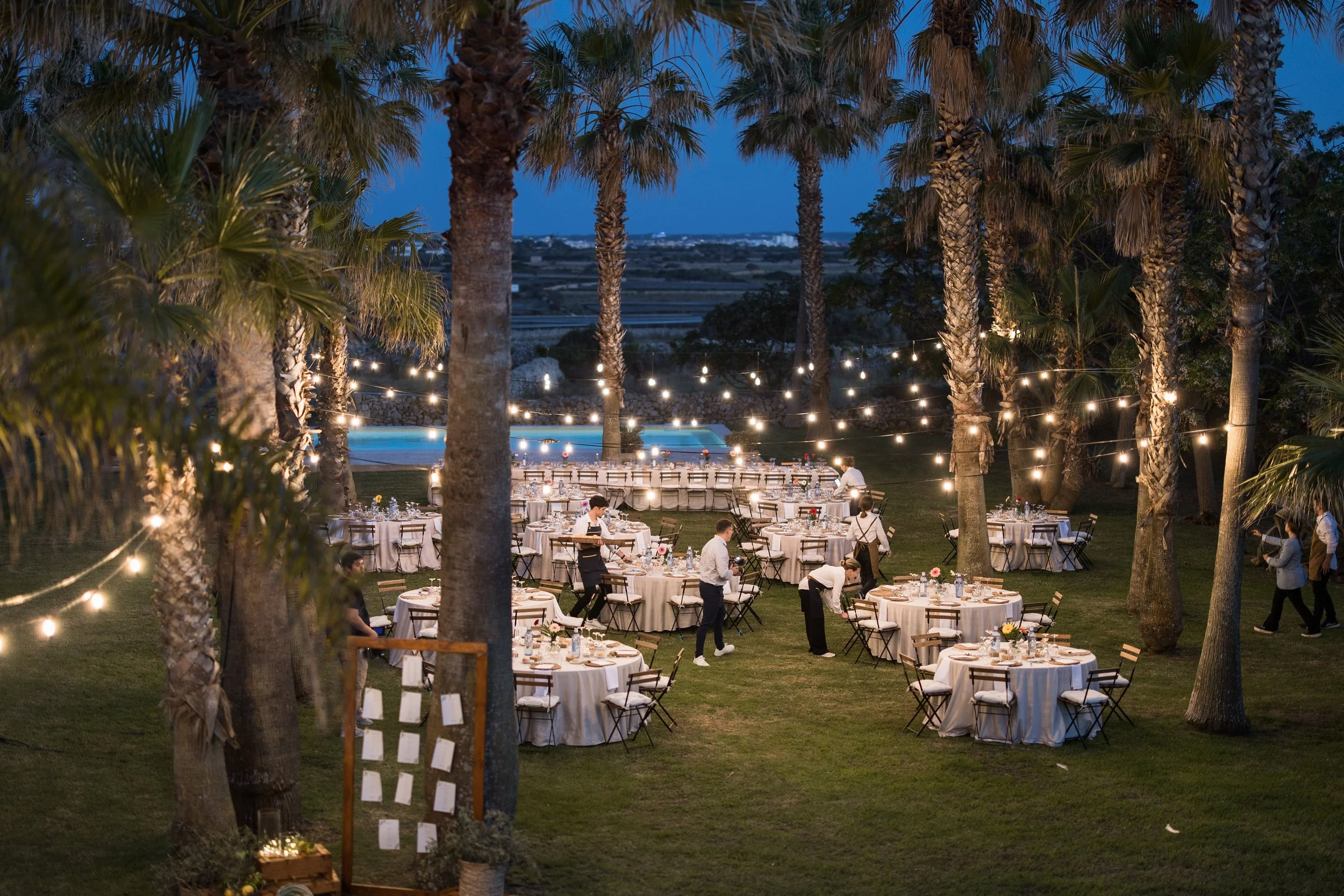 Cena al aire libre con mesas redondas decoradas con manteles blancos, en un espacio rodeado de palmeras y luces suspendidas, en un entorno nocturno.