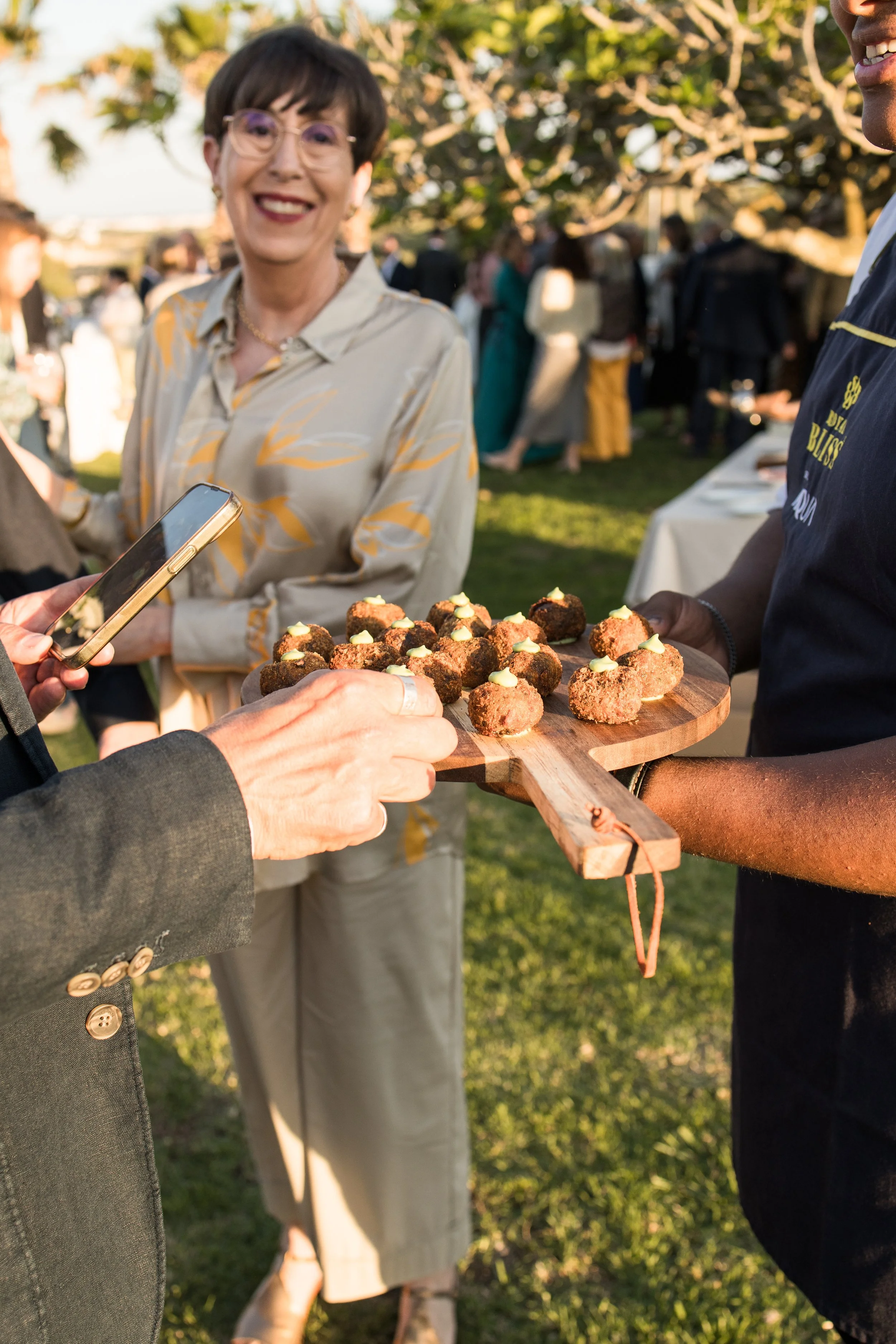 Persona sirviendo comida en un evento al aire libre, con una mujer sonriendo en el fondo y varias personas en el fondo socializando y disfrutando de la reunión.