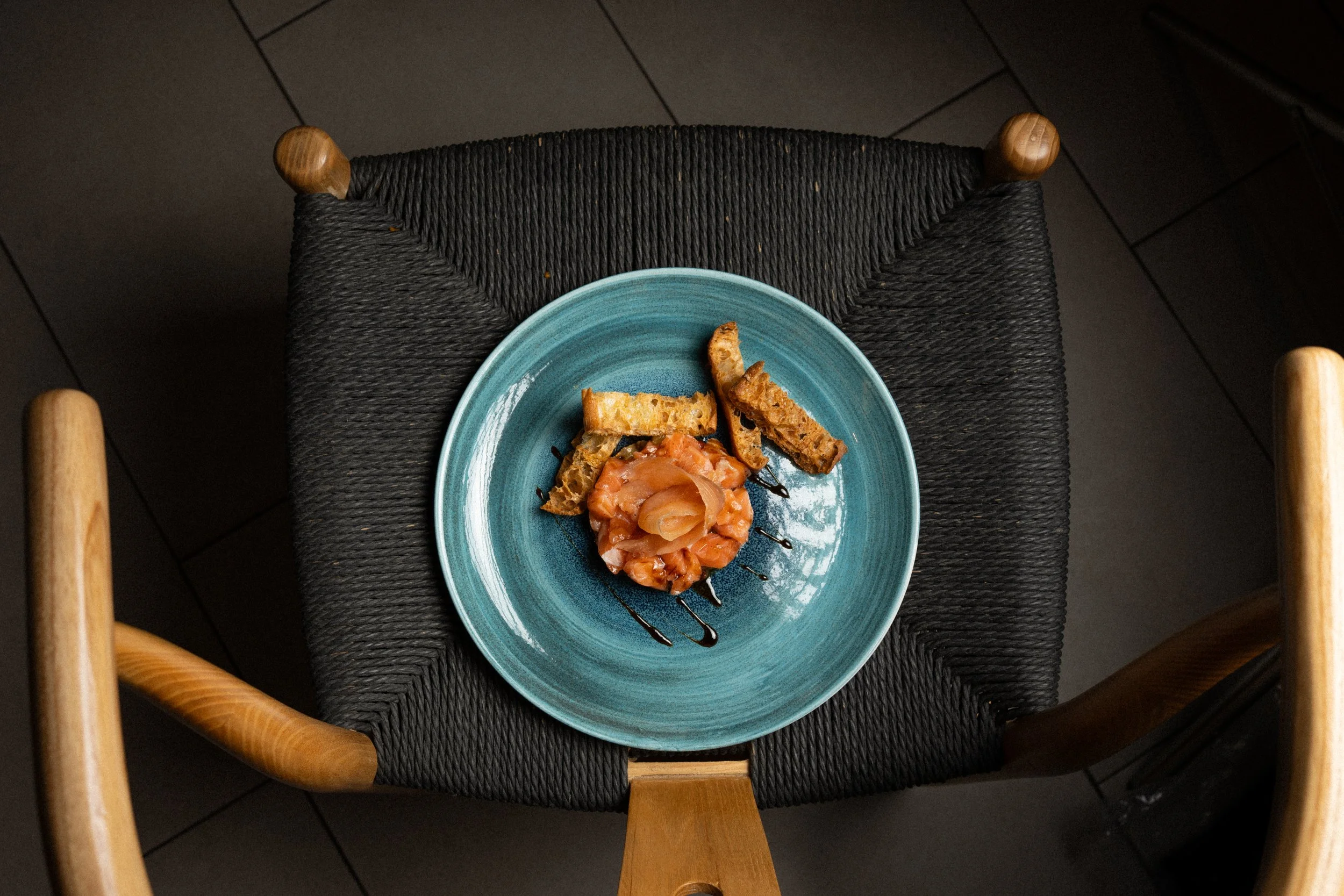 Plato con sashimi de salmón, acompañado de pan tostado y un chorrito de salsa de soja, sobre un plato azul en una silla de madera con asiento de cuerda negra.