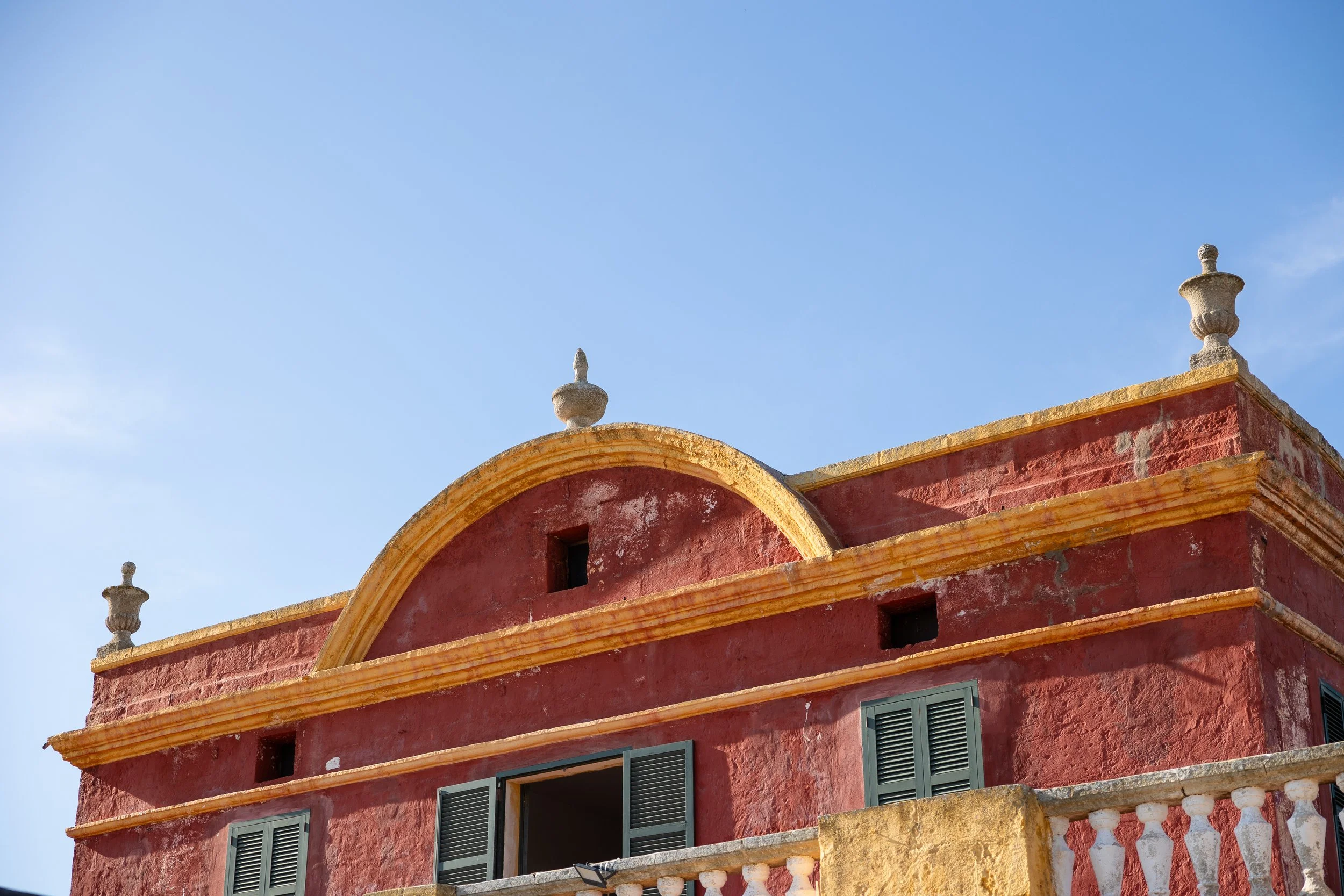 Edificio antiguo de color rojo con detalles en amarillo y ventanas con persianas verdes, bajo un cielo azul claro.