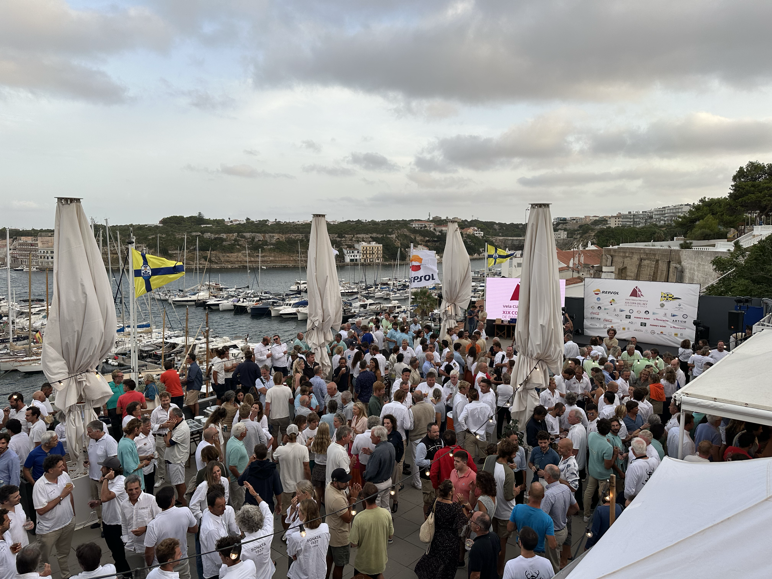 Gran concurrencia de personas en un evento junto a un puerto deportivo con barcos amarrados, banderas de diferentes colores y un escenario con pantallas y cartel con logotipos.