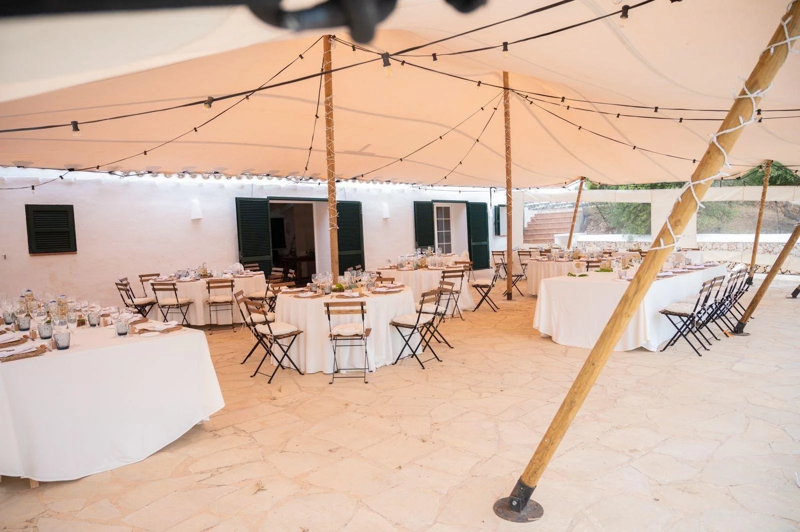 Decoración de evento al aire libre con mesas redondas con mantelería blanca, sillas de madera, vajilla, copas y botellas de agua, cubiertas por una carpa grande con luces colgantes, en un entorno rústico con paredes blancas y ventanas con persianas v