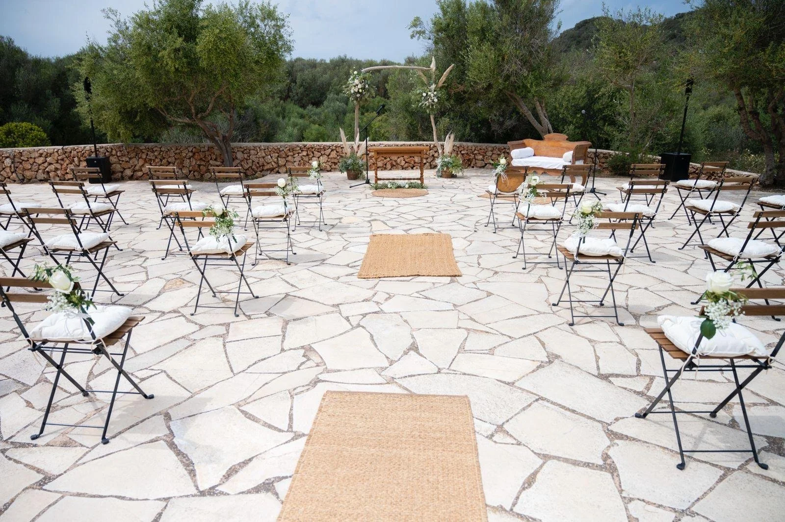 Área de ceremonia al aire libre decorada con sillas de metal con cojines blancos, camino de yute y un arco nupcial con flores. Jardín en el fondo con árboles y montañas.